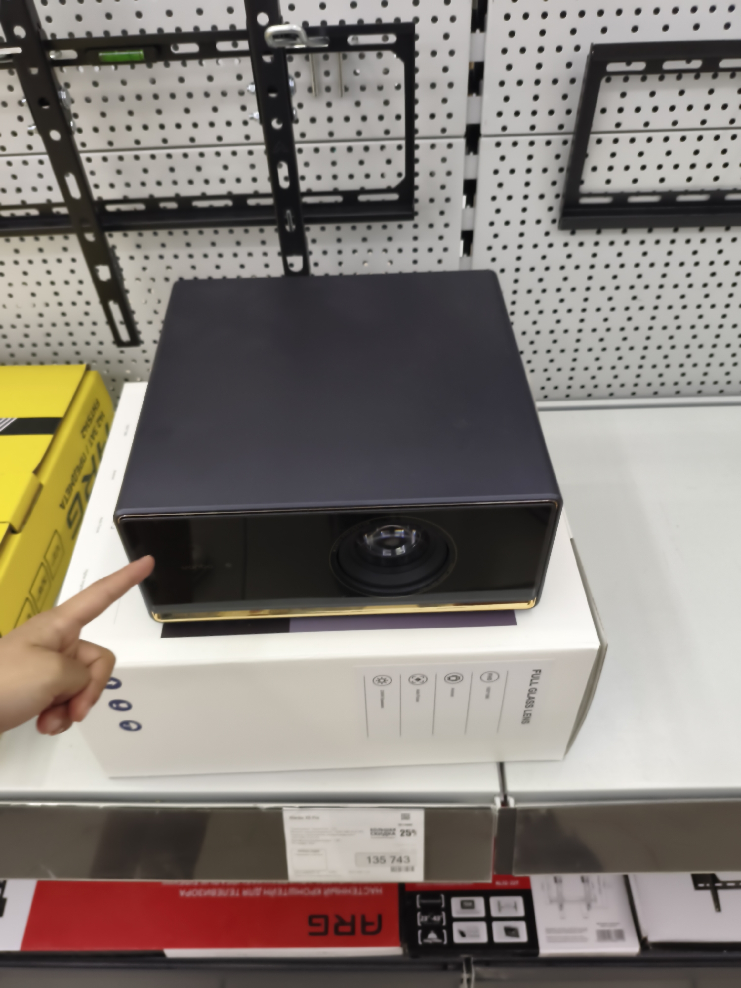 A person's left hand is pointing at a dark-colored projector box sitting on top of a white box. The projector box has a gold trim around the lens. The white box has some symbols and text in black, including "FULL GLASS LENS." To the left of the projector box is a yellow box with black text. In the background, a pegboard with black mounting brackets is visible. The overall scene appears to be a retail environment, likely a store in Aktobe, Kazakhstan, as suggested by potential context related to products commonly sold in such locations in that region.