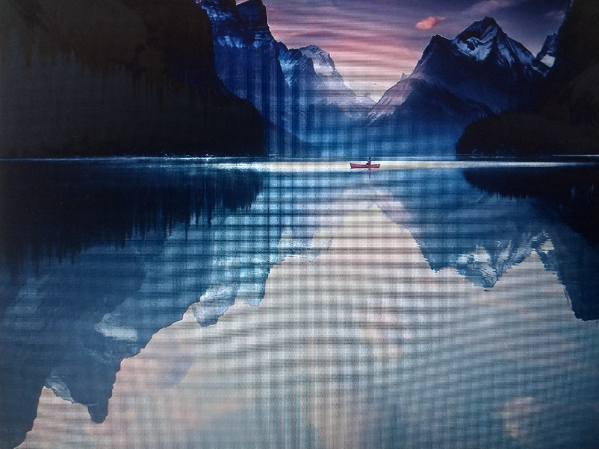In an expansive, still lake that mirrors the surrounding mountainous landscape, a lone individual is seated in a red canoe. The canoe is positioned in the center of the frame, directly on the horizon line where the water meets the sky. The sky is painted with soft hues of pink and purple, suggesting either dawn or dusk, transitioning to deep blues as it ascends towards the jagged peaks of the mountains. These mountains, majestic and imposing, are covered in snow on their upper reaches, creating a stark contrast with their dark, forested lower slopes. The reflection of the mountains and the sky is perfectly rendered on the glassy surface of the lake, creating a symmetrical and serene scene. The overall atmosphere is one of tranquility and isolation, with the solitary figure in the canoe emphasizing the vastness and solitude of the natural environment.