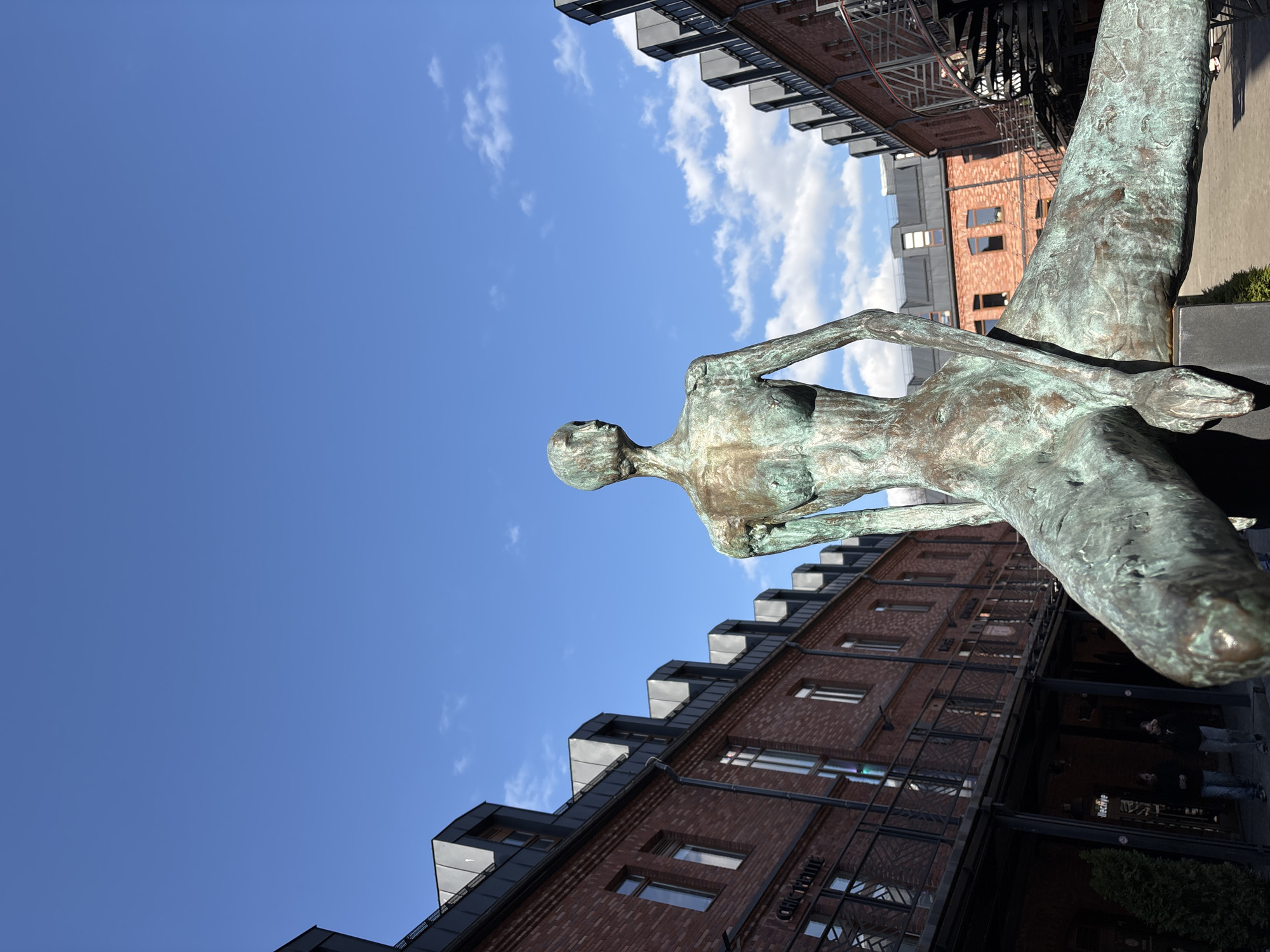The image depicts a contemporary bronze statue of a stylized, slender female figure with an elongated lower body, suggestive of a mermaid or a reclining form, positioned centrally. The figure exhibits significant verdigris, indicating aging or intentional patination, and faces towards the upper right of the frame. Her head is bald and her features are minimalist; her torso is bare, and her arms are either absent or indistinguishably merged with her form. She appears to be seated on a dark, rectangular base.
The scene is set in an urban environment, specifically within a courtyard or passage flanked by two multi-story red-brick buildings. These structures feature dark grey pitched roofs with numerous modern dormer windows. The building on the left shows signage in Cyrillic, including "СИНОПСИС" and "КОНЦЕПТУР," and has a lower level with windows and black railings. The building on the right presents similar architectural characteristics and includes a prominent black spiral staircase on its facade.
The ground surface is a dark paved area. In the lower left, two individuals are walking away from the viewer, dressed in dark jackets and jeans. They are positioned near the base of the left building, which also features some green shrubbery. The sky above is clear and bright blue with scattered white cumulus clouds, indicating sunny weather.
This location is identified as New Holland Island (Новая Голландия) in Saint Petersburg, Russia, known for its renovated industrial architecture and public art installations. The statue is a permanent feature within this cultural and recreational complex.