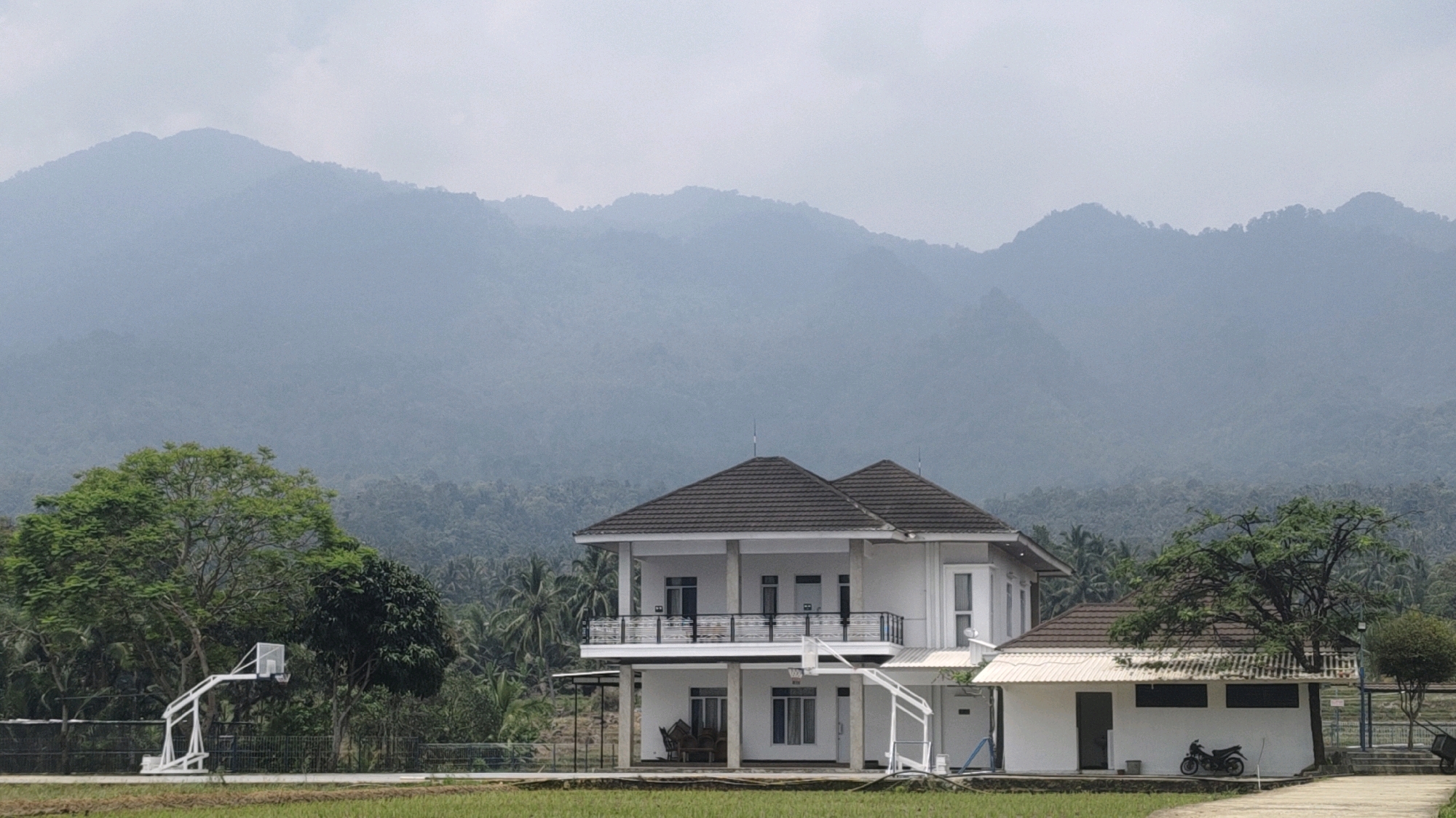The image depicts a serene landscape in Ujungtebu, Indonesia, dominated by a large, two-story white building with a dark tiled roof and balconies. To the right of this main structure is a smaller, single-story white building with a similar roof, featuring a motorcycle parked near its entrance. The foreground is a lush green field, possibly rice paddies, with a basketball hoop and backboard visible on the left side of the frame, suggesting recreational facilities. In the background, dense tropical foliage, including palm trees, blankets the rolling hills leading up to majestic, mist-shrouded mountains. The overall atmosphere is tranquil and natural, with the buildings integrated into the scenic environment.