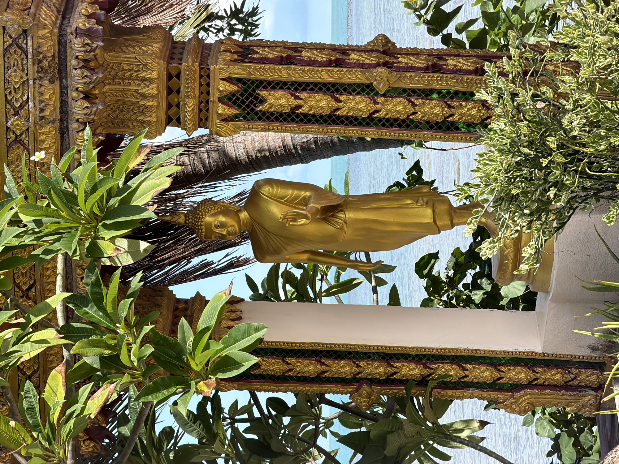 A golden statue of Buddha stands within an ornate, gilded pavilion in Baan Plai Laem, Thailand. The statue is positioned centrally, facing forward, with its right hand raised in a gesture of blessing or protection. The pavilion features intricately carved golden pillars with decorative elements in red and green. Lush green foliage, likely tropical plants, surrounds the statue and the pavilion, with some leaves partially obscuring the view. In the background, the clear blue water of the ocean stretches to the horizon, under a bright blue sky. A palm tree trunk is visible behind the statue, suggesting a coastal or island setting. The overall scene evokes a sense of peace and tranquility, typical of religious sites in Southeast Asia.