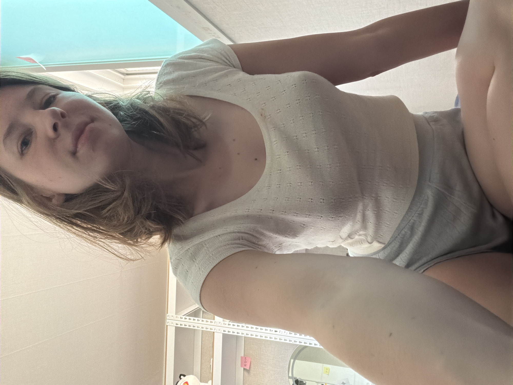 An individual is seated in a room, with their left arm extended toward the camera. The subject is wearing a white, short-sleeved, scoop-neck top and gray shorts. They have light brown hair. The room appears to be a bedroom or living space, with a ceiling that has horizontal paneling. To the left of the subject, there is a white shelving unit with various items on it, including what appears to be stuffed animals and a white electronic device. A small red sign with Korean writing is affixed to the shelving unit. To the right of the subject, a window with a bright blue tint is visible, and a white vertical blind is partially obscuring the view. The flooring is not clearly visible. The lighting in the room is soft and appears to be natural light coming from the window. The image was taken in Dongducheon-si, South Korea.