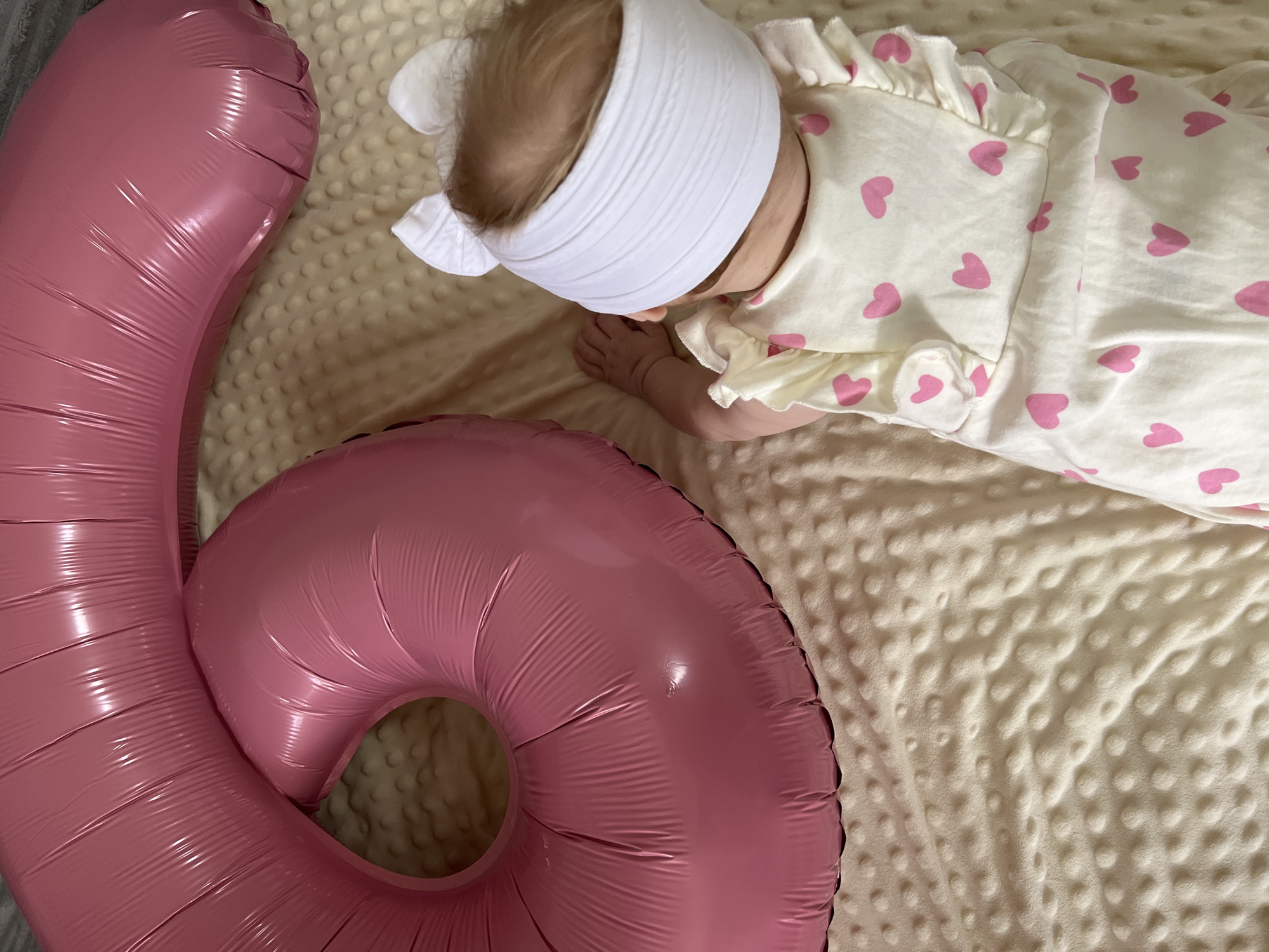 A baby lies on a beige blanket next to a pink number six balloon. The baby wears a white headband with cat ears and a white onesie with pink hearts. The location is Chaikovsky, Russia.