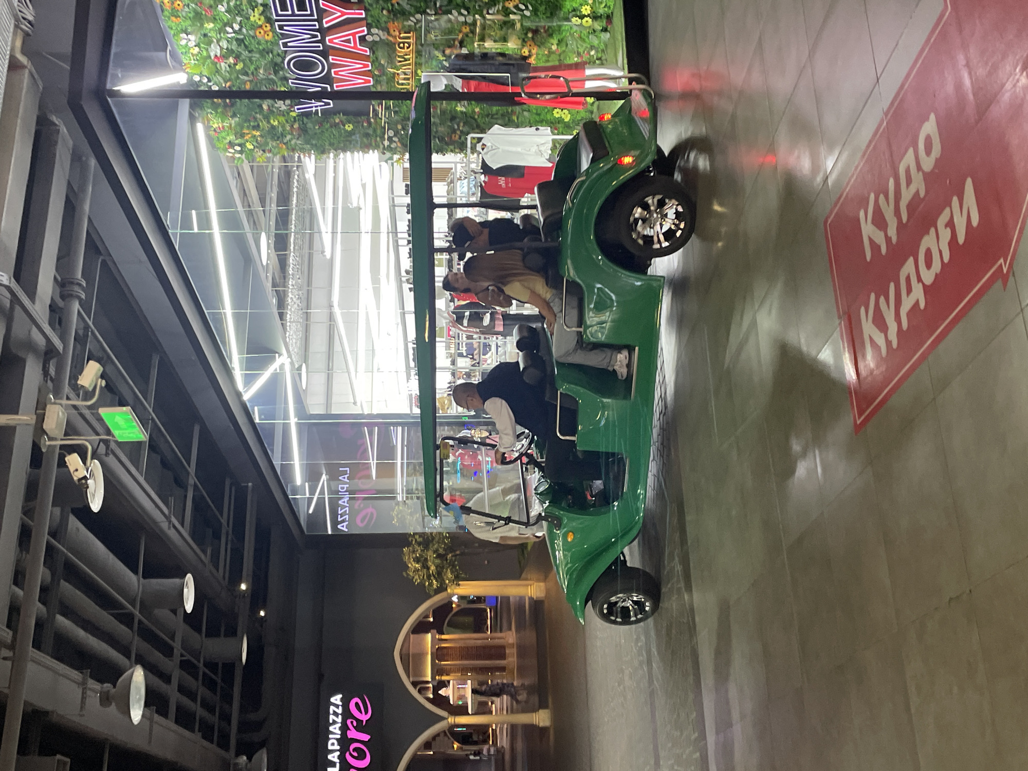 An outdoor pedestrian zone in Almaty, Kazakhstan, features a green golf cart with four occupants. The cart is positioned in the foreground, traveling from left to right. The driver, an older man wearing glasses and a white shirt with a dark vest, is at the wheel. Behind him, two women are seated, and a third person, whose face is obscured, is also in the cart.
To the right of the golf cart is the storefront of a shop with a large glass window displaying mannequins dressed in clothing, some of which is red. A prominent neon sign above the display reads "WOMEN WAY" in white letters with a red outline, and below it, "dm wen" in white. The storefront is adorned with a vertical arrangement of artificial flowers and greenery.
To the left of the golf cart, a large building with black exterior walls is visible. Above this building, a complex network of pipes and lighting fixtures is suspended from the ceiling, suggesting an indoor or covered outdoor space. A pink neon sign on the black wall reads "LA PIAZZA". Further to the left, a structure with arched entrances and decorative columns is visible.
On the tiled ground in the foreground, a red sign with white Cyrillic lettering is laid out in an arrow shape. The visible portion of the text appears to be "куда куда" and "кудаофи". The overall lighting suggests it is either evening or a dimly lit indoor area.
