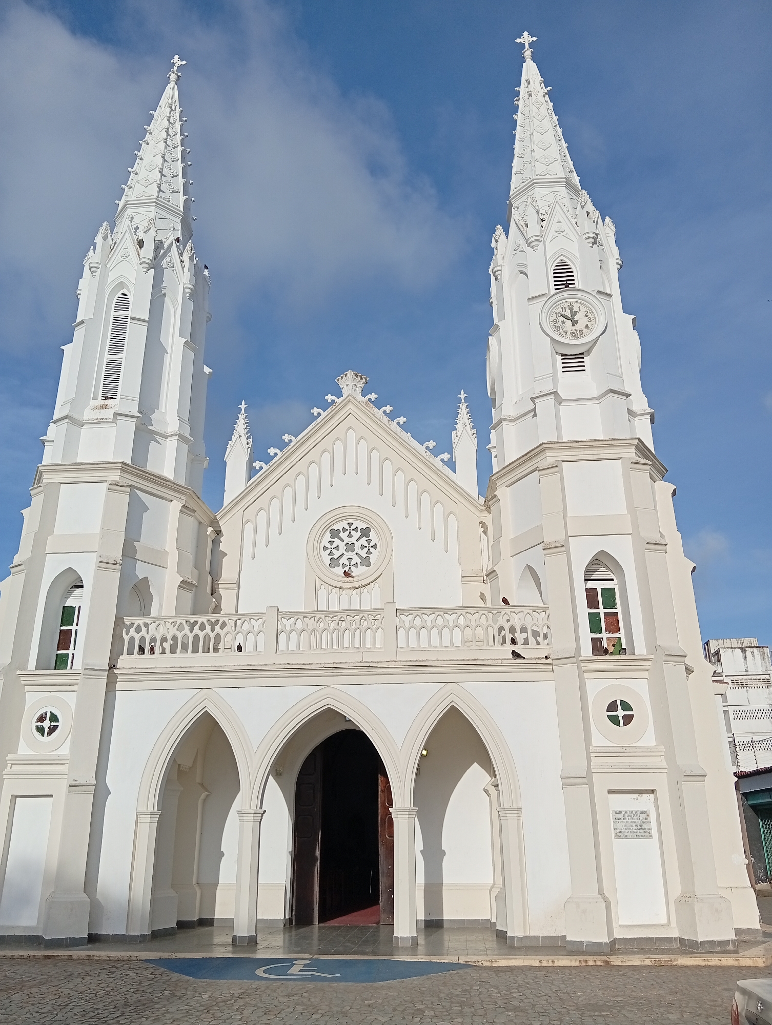 A white, neo-gothic style church with two prominent spires and a clock tower stands against a clear blue sky with scattered clouds. The church features arched doorways, a decorative balcony, and stained-glass windows. Pigeons are visible on the balcony railing and near a window. The ground in front of the church is paved with cobblestones, and a blue square with a white accessibility symbol is painted on the ground, indicating a designated parking spot for individuals with disabilities. The scene depicts the Church of Our Lady of the Valley (Iglesia de Nuestra Señora del Valle) in Juan Griego, Venezuela.
