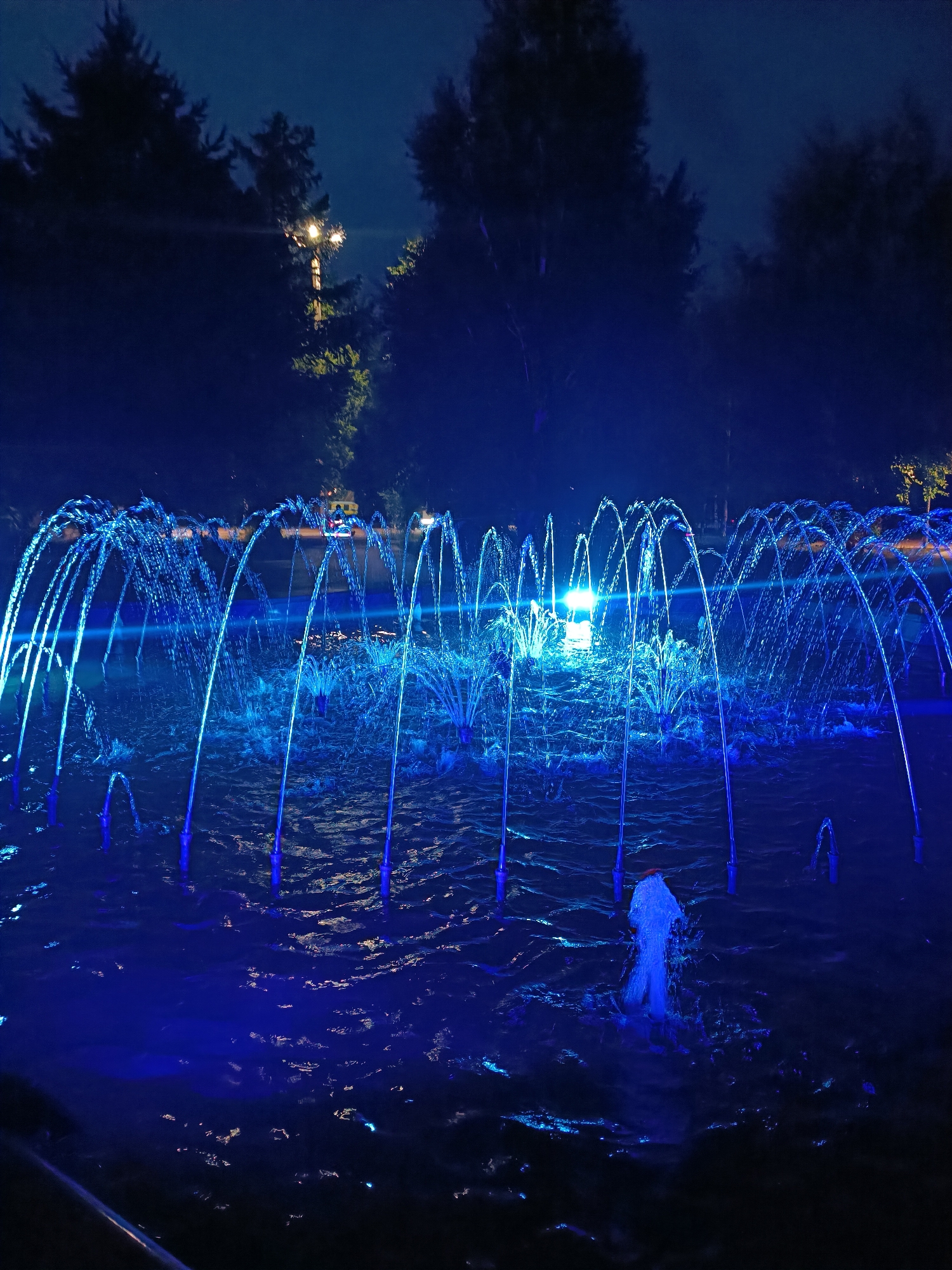 The image captures a fountain in Kokshetau, Kazakhstan, at night. The fountain is illuminated with blue lights, creating a striking visual effect. The water jets arch gracefully, forming a symmetrical pattern. The surrounding environment is dark, with silhouettes of trees visible in the background. A street lamp is also visible in the background. The water surface is slightly rippled.