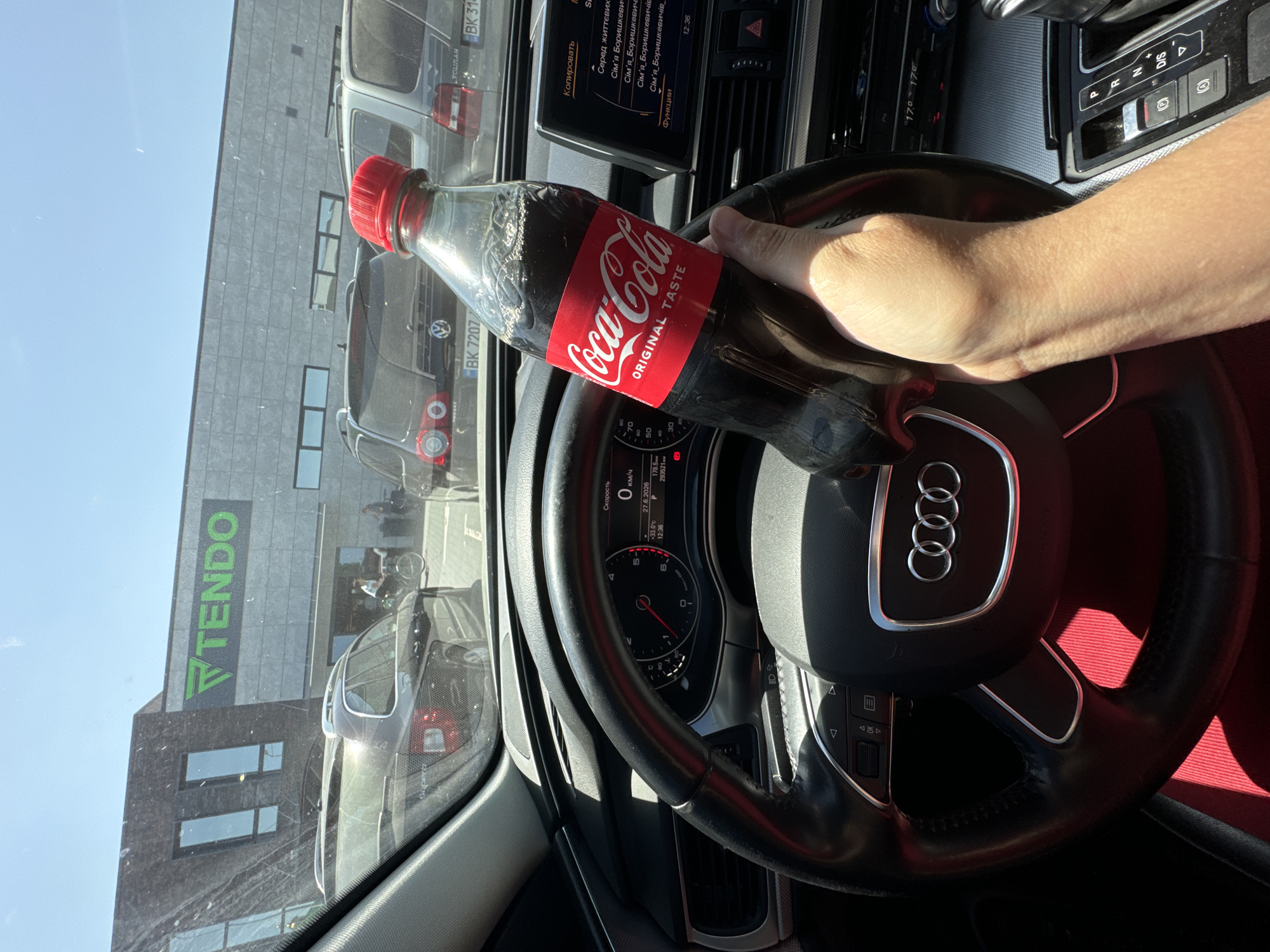 A first-person perspective shot from inside a car shows a hand holding a Coca-Cola bottle. The car's dashboard and steering wheel are visible. The background shows a building with a large "TENDO" sign, suggesting a commercial establishment. Several cars are parked outside, and a person is seen on a bicycle. The scene is set in Sarny, Ukraine.