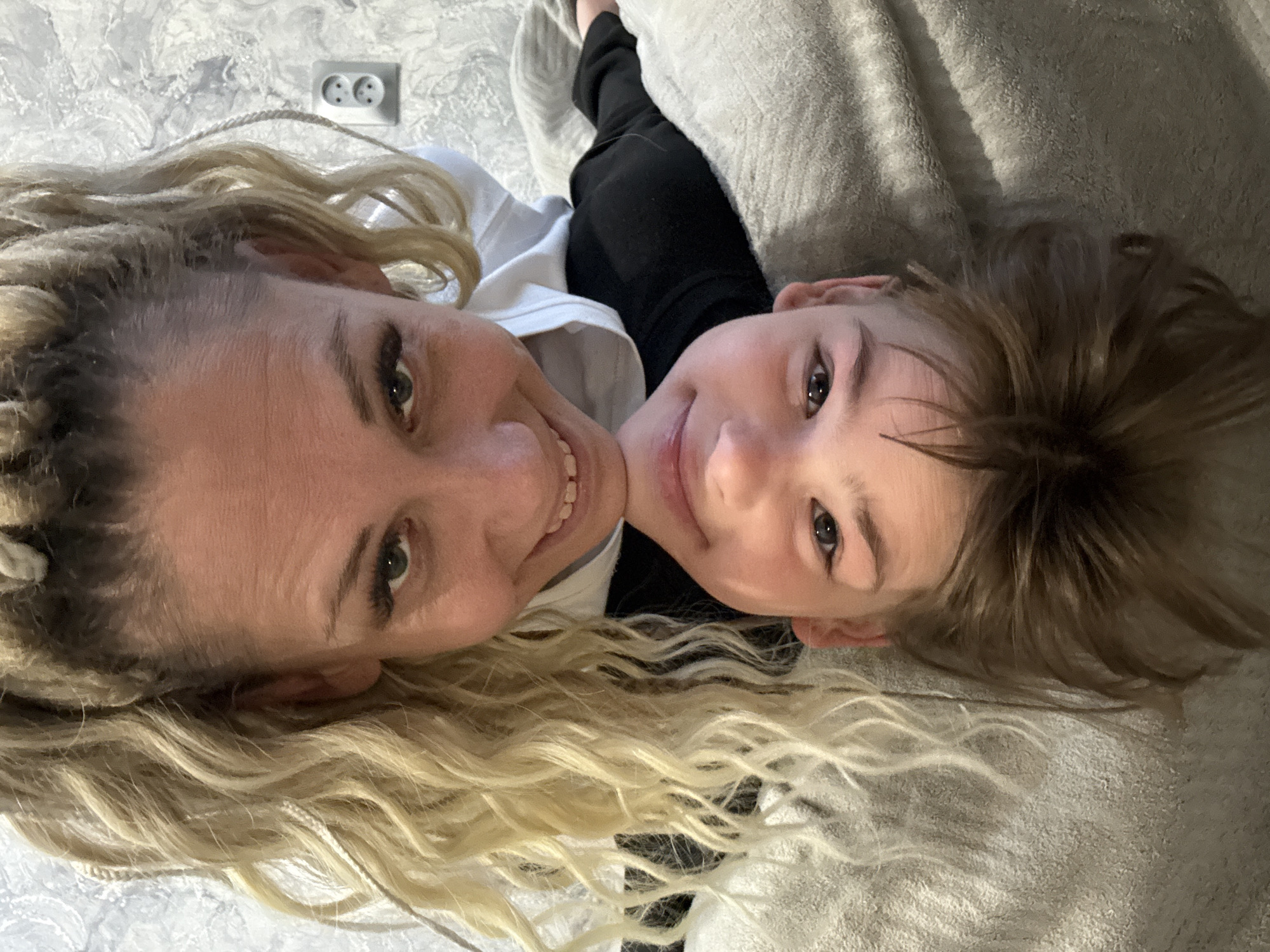 A selfie-style photograph captures two individuals, a woman and a child, lying close together. The woman, on the left, has long blonde hair styled with braids. Her face is visible, displaying a smile, and her eyes are open, looking towards the camera. To her right, a child with short brown hair also smiles and looks at the camera. They are wearing a white and black top. In the background, there is a gray blanket and a partially visible electrical outlet on the wall. The photo appears to have been taken indoors. There is no information to conclude the location in the city of Rusanovo, Russia.