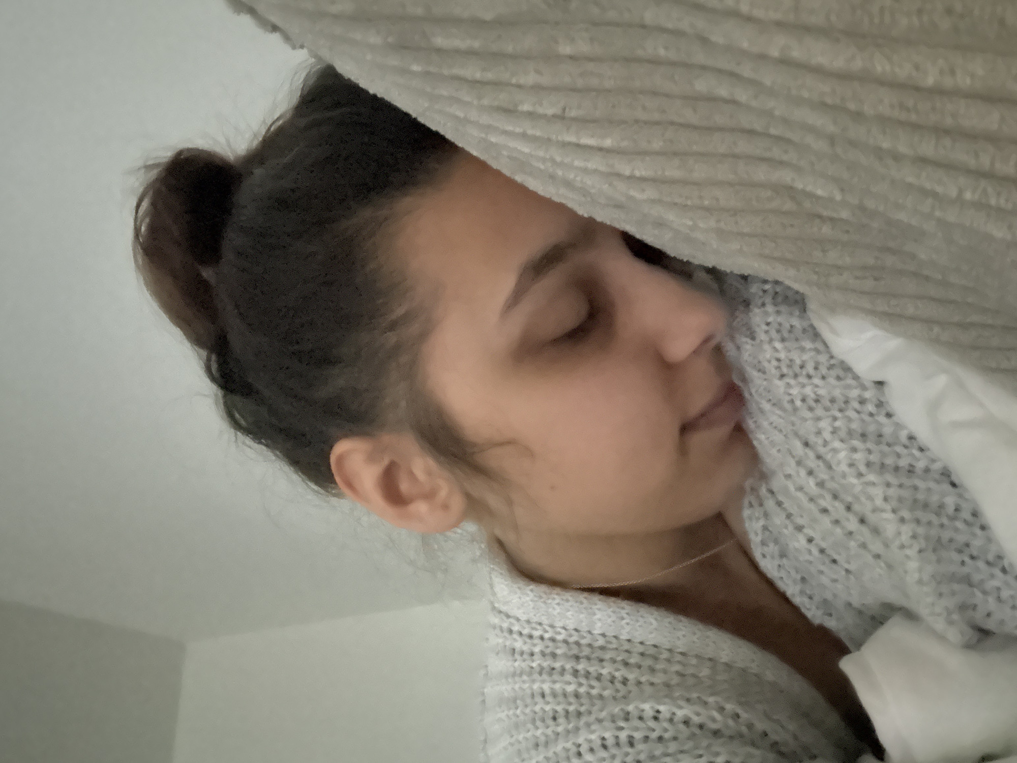 A woman with dark hair pulled back in a bun is seen sleeping, partially covered by a gray knitted blanket and a white sheet. Her eyes are closed, and her face is relaxed. The background consists of a light gray wall. The location is not discernible from the image and cannot be determined to be Mönchengladbach, Germany.