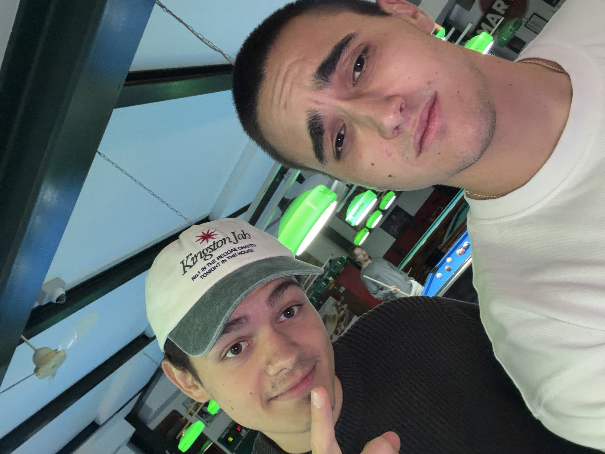 Two young men are posing for a selfie. The man in the foreground is wearing a hat with the words "Kingston Jah" on it. In the background, another man is playing pool. The environment appears to be a pub or bar with pool tables. The lighting is provided by hanging green lamps. Based on the OCR text in the background, the location might be in Milan, Italy.