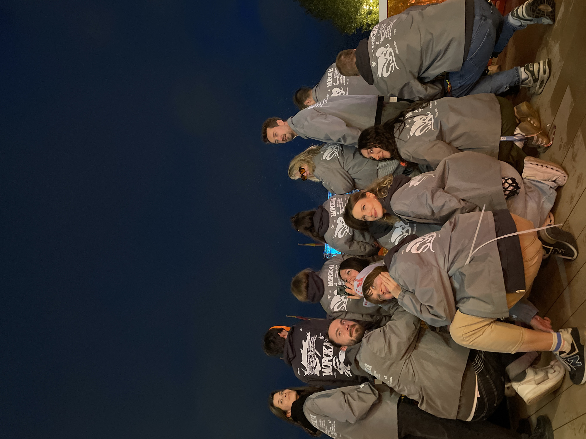 A group of people in gray jackets with a logo featuring the word "MOPCKAR" and a graphic are posing on a wooden surface, likely a boardwalk or deck. The background shows a dark blue sky suggesting it is nighttime. Some of the individuals are smiling or looking directly at the camera. The location is likely in Saint Petersburg, Russia, based on the lettering style on the jackets.