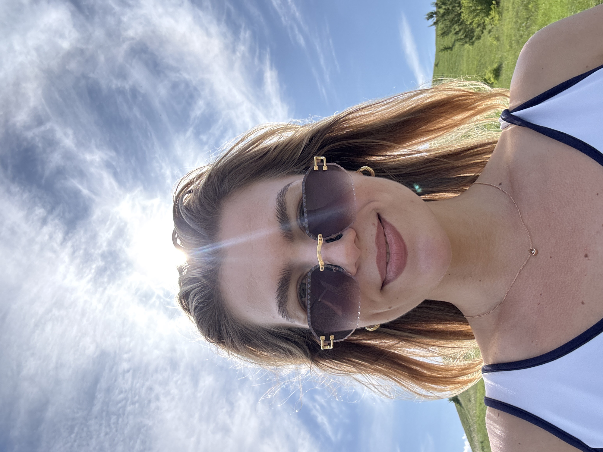 A close-up outdoor selfie depicts a smiling female with long, light brown hair, wearing sunglasses. Her head is positioned centrally, with bright sunlight originating from behind and above, causing a prominent lens flare across the upper portion of the image and backlighting her hair. She wears large, rimless dark sunglasses with gold-colored accents, small gold hoop earrings, and a delicate gold-colored chain necklace with a small teardrop pendant resting on her sternum. Her lips are closed, showing a subtle smile with visible dimples on her cheeks. She is wearing a white tank top with dark-colored trim on the straps. The background consists of a clear blue sky with scattered wispy clouds. Below the sky, green grassy hills are visible, with some small dark green bushes or trees on the right. The location is in the city of Unknown, Unknown.