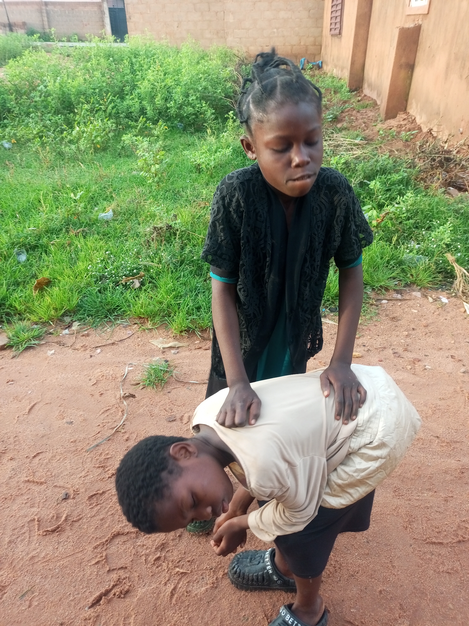 Two young children are interacting in an outdoor environment. The child in the foreground, wearing a light-colored shirt and dark shorts, is bent over with their head down. The child standing behind them, wearing a black lace top over a green garment, has their hands placed on the back of the bent-over child. The ground is sandy and reddish-brown, with some sparse green vegetation and dry leaves scattered around. In the background, a tan-colored building with a window and a dark-colored door is visible, surrounded by more greenery. The scene appears to be in a rural or semi-urban area, possibly within Tempure, Nigeria, based on the architectural style and vegetation.