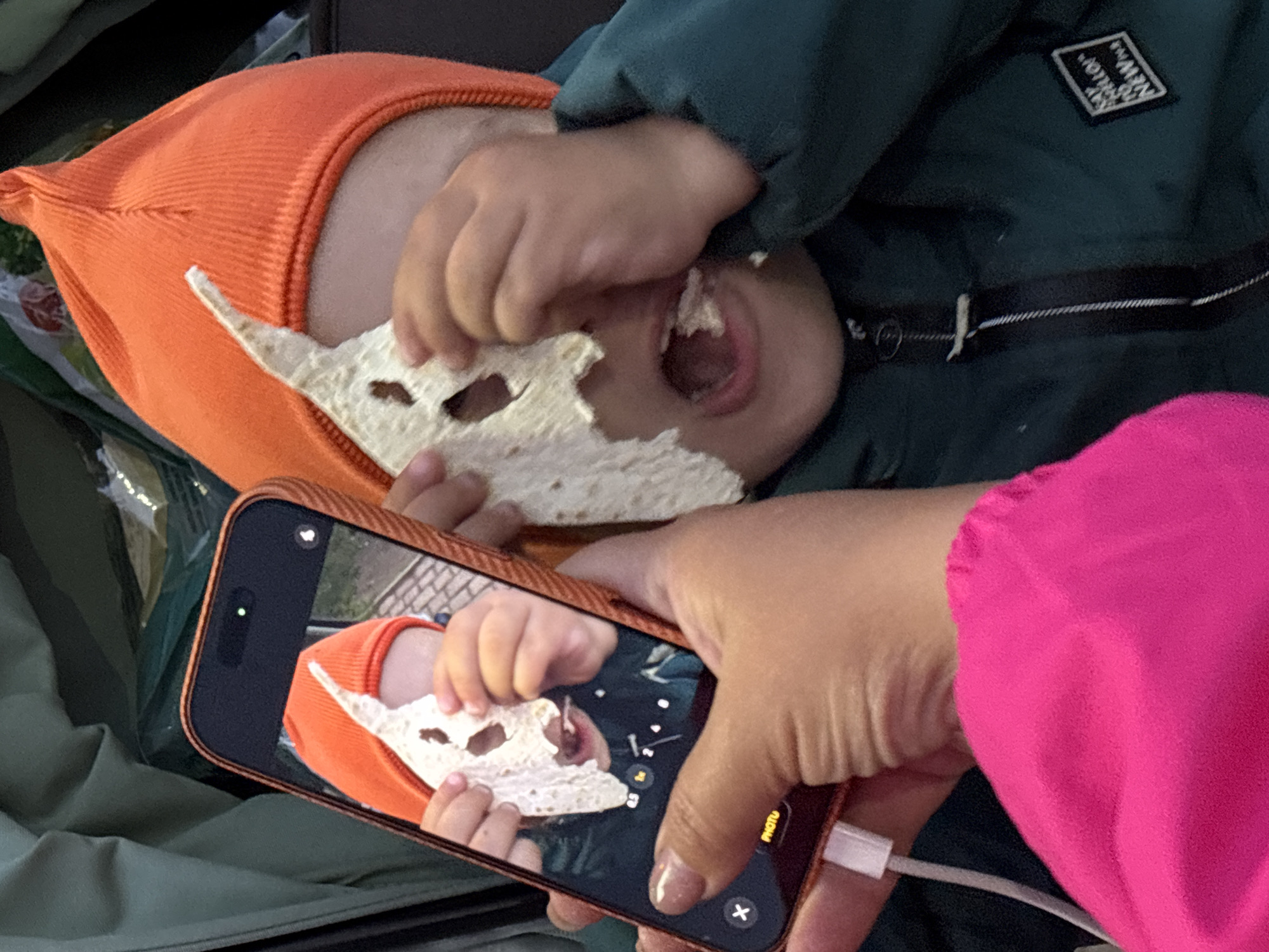 The image depicts a close-up scene occurring indoors, in the city of Chaikovsky, Russia. A child, wearing an orange ribbed knit hat and a dark green zippered jacket with a partially legible white patch on the right chest ("SAY HELLO TO NEW"), is visible in the foreground. The child's left hand holds a piece of irregularly shaped, thin, light-colored flatbread to their face, obscuring their eyes and nose. This flatbread has several torn or cut holes, giving it the appearance of a rudimentary mask. The child's mouth is open, revealing a piece of food inside, suggesting they are actively eating.
To the left, an adult's hand, featuring manicured nails with a light polish, holds a black smartphone (identifiable as an iPhone with a Dynamic Island notch) encased in an orange protective cover. A white charging cable is connected to the bottom of the phone. The phone's screen displays a live view from its camera, capturing the child in a similar pose with the flatbread mask. The adult's wrist and forearm are partially visible, covered by bright pink fabric. A second piece of flatbread is partially visible on the far right, near the child's right shoulder. The background is dark and indistinct, with hints of a green/khaki fabric on the upper left, suggesting an enclosed space such as a vehicle interior or a room.
