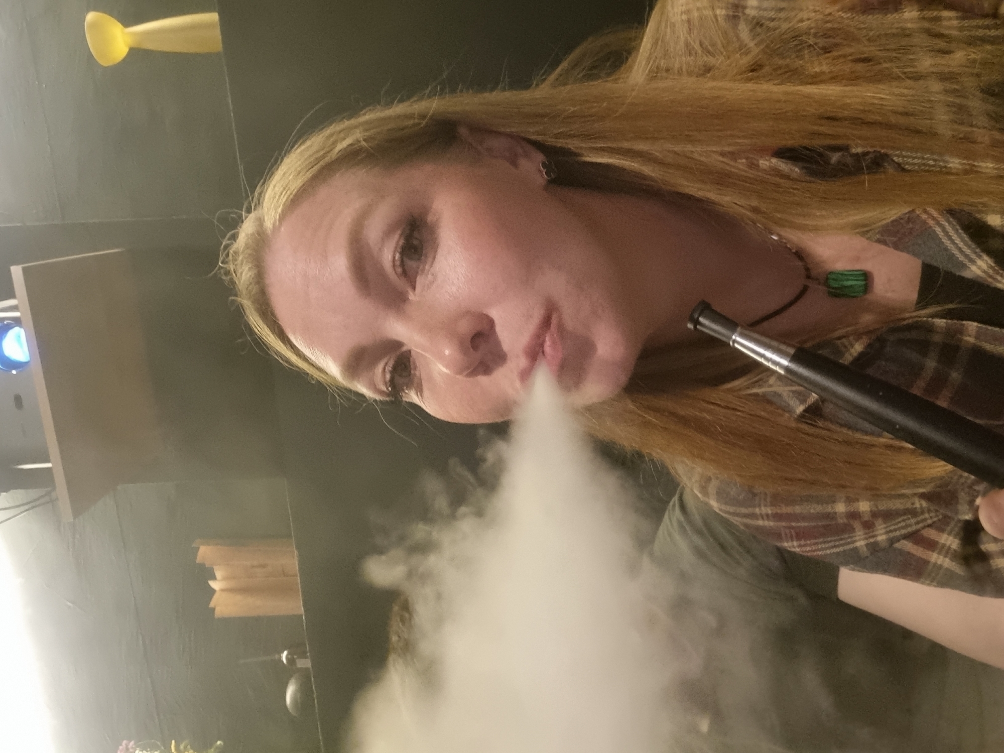 The image displays an angled, medium-close-up shot of a woman exhaling a thick cloud of vapor or smoke from a dark, sleek device, likely a vape pen or a part of a hookah. She has long, straight blonde or light brown hair and looks towards the viewer with a calm expression. She is wearing a plaid shirt, predominantly in shades of brown and red, over a dark undershirt. A black cord necklace with a distinctive rectangular green pendant, featuring striped patterns, hangs around her neck. A small, dark earring is visible in her left ear.
The setting is an indoors, dimly lit environment, suggesting evening or nighttime. The background is dark, with various subtle elements. In the upper left, a bright yellow, elongated object protrudes from a shelf. Below it, a subtle blue glow emanates from a device or light source on a lighter surface. Further into the background, a stack of light-colored, possibly wooden or paper objects, is visible on another shelf. The overall atmosphere implies a relaxed social setting, such as a lounge or cafe, in Chaikovsky, Russia. There is no visible text in the image.