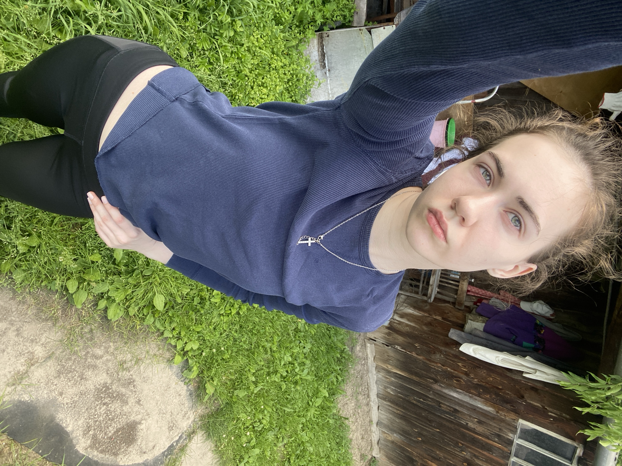 The image shows a young woman with light skin, blue eyes, and light brown hair, taking a selfie outdoors. She is wearing a navy blue long-sleeved shirt and black leggings. A silver necklace with a cross pendant is visible around her neck. Her facial expression is neutral, and she appears to be looking directly at the camera.
The setting is an outdoor area with lush green grass and some dirt patches. In the background, there is a rustic wooden structure, possibly a shed or an old building, with wooden planks. Various items are piled up inside or near the structure, including what appears to be folded laundry in different colors like purple, grey, and white, and a green bucket. A small window is visible on the wooden structure.
The lighting suggests it is daytime, and the greenery indicates a season with favorable weather. The overall scene appears to be in a rural or semi-rural environment. There is no visible text in the image. The woman's posture and the way she is holding the camera suggest she is standing or crouching. The image is oriented vertically.
