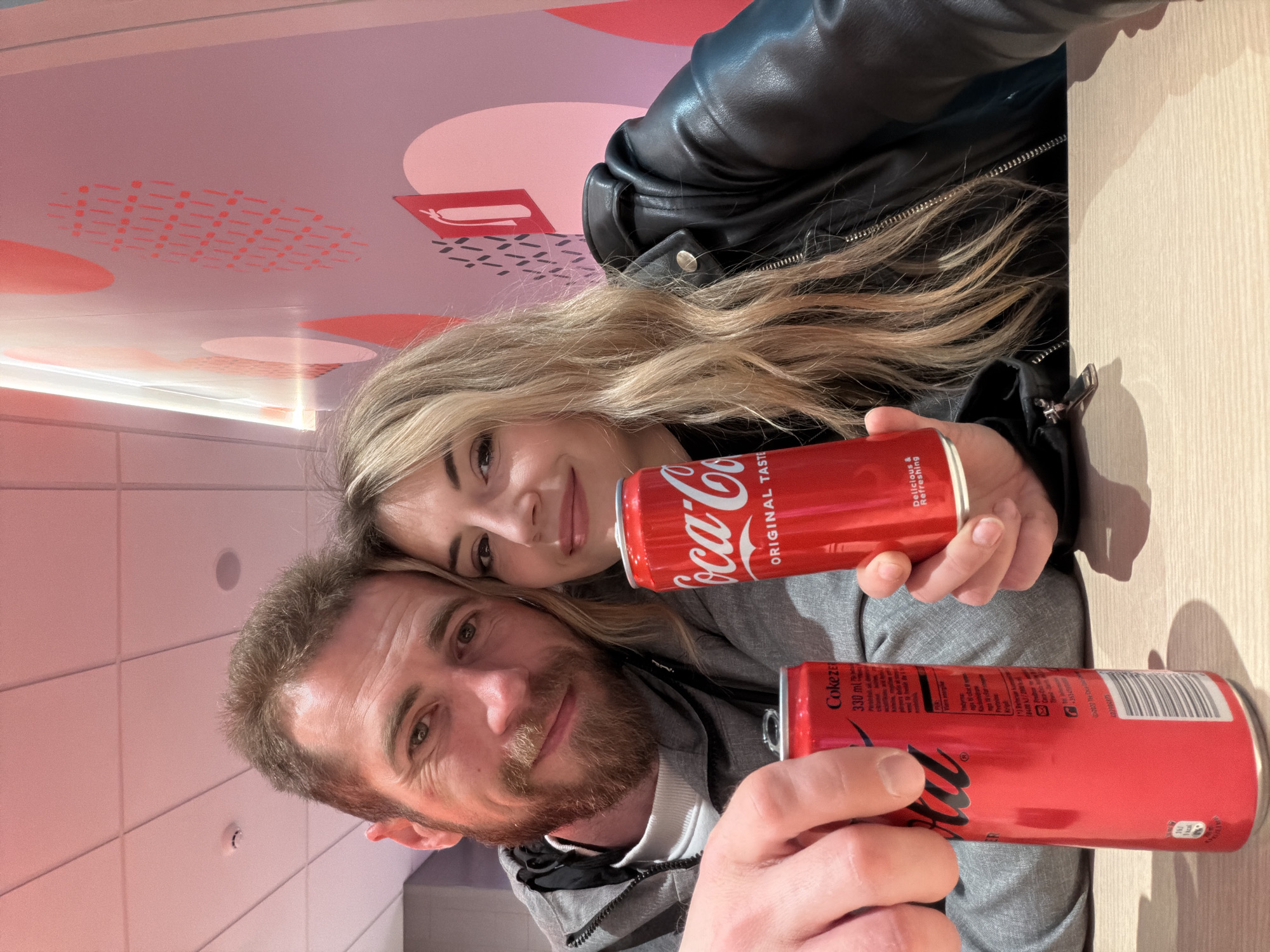 A man and a woman are holding two Coca-Cola cans in an indoor setting with a modern design. The man, with a beard and wearing a grey hoodie, is situated on the lower left of the frame. The woman, with blonde hair and wearing a black leather jacket, is positioned above him. The background features a light-colored wall and a ceiling with pink and white circular and rectangular designs. Based on the style and ambiance, it is plausible that the location is a restaurant or cafe in Budva, Montenegro.