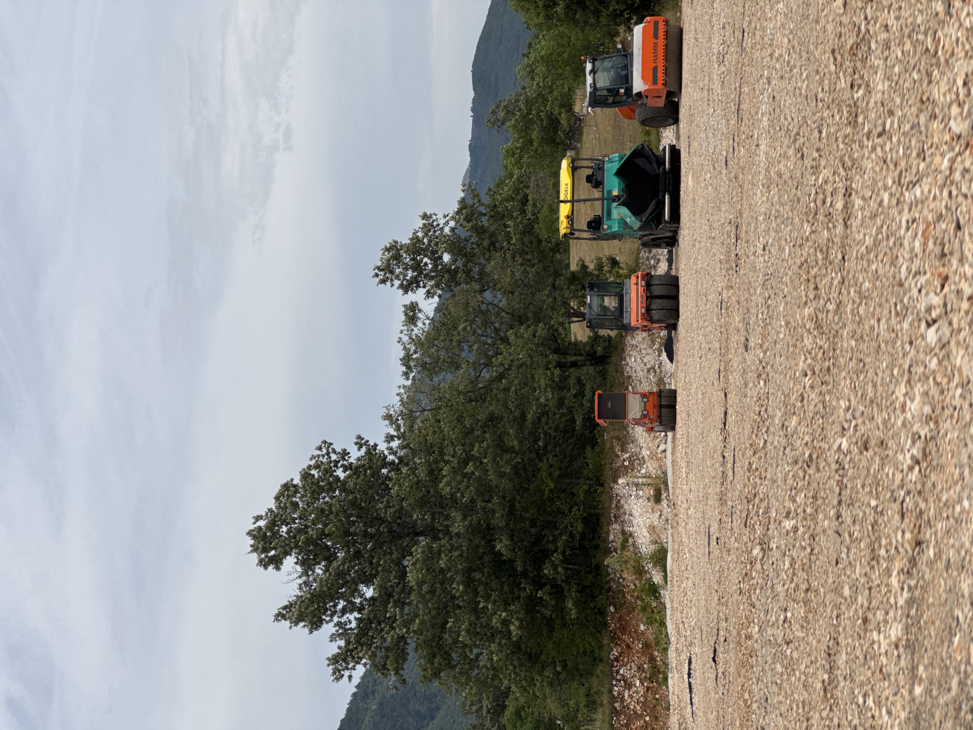The image depicts a construction site in Jasenovo Polje, Montenegro. The foreground is a vast expanse of gravel. In the middle ground, there are three pieces of heavy machinery: two compactors and one asphalt paver. The compactor on the left is orange and black, the asphalt paver in the center is green and yellow, and the compactor on the right is orange with black accents. Behind the machinery, a dense, dark green forest with a large, mature tree dominates the scene. Further in the background, rolling hills covered in vegetation are visible under a cloudy sky. The overall impression is one of an active construction project in a natural, rural setting. There are no people visible in the image.