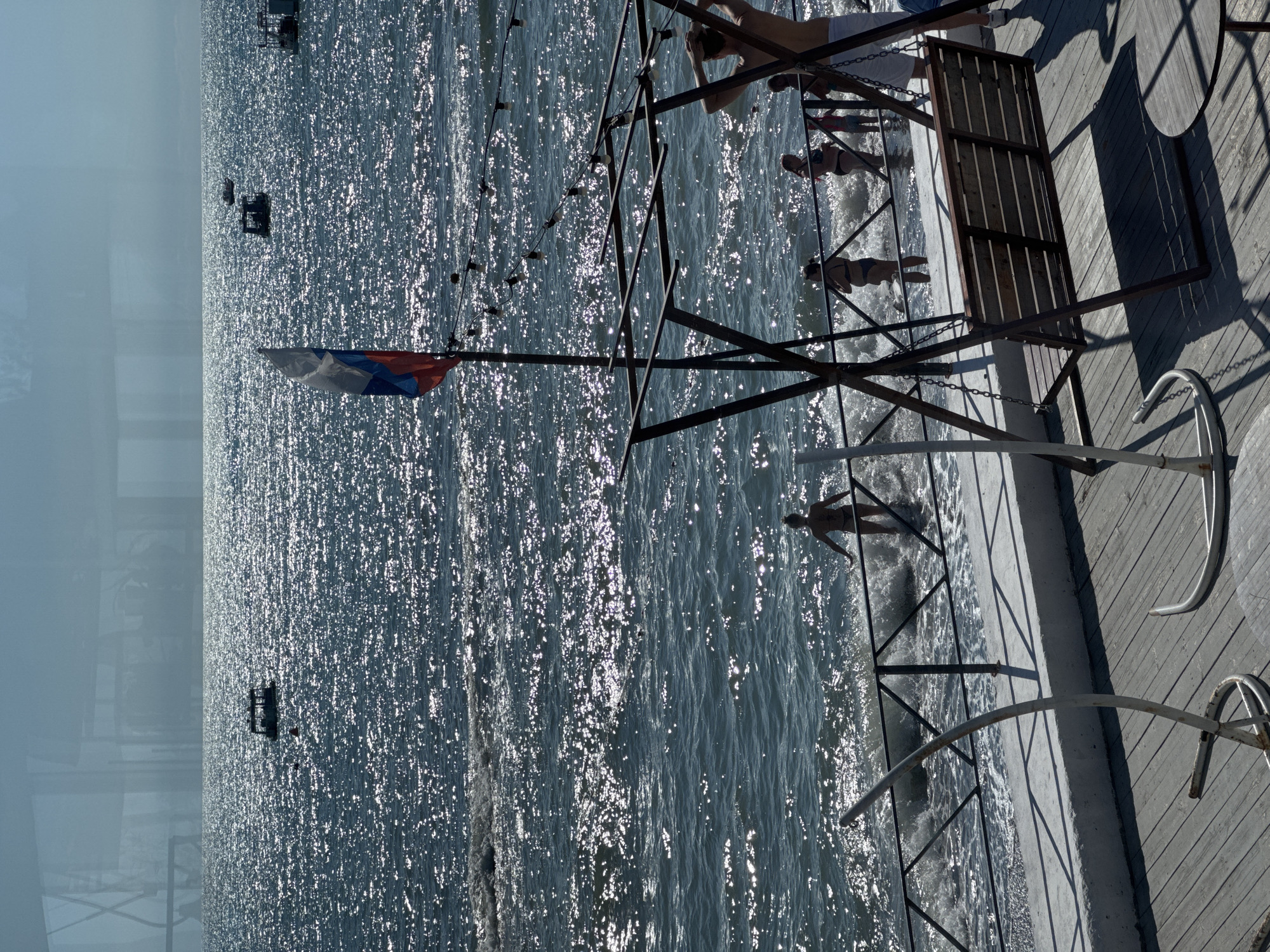 The image depicts a sunny day at a beach in Sochi, Russia. The sea is sparkling with sunlight, and several boats are visible in the distance. In the foreground, a Russian flag waves on a flagpole. A metal swing set and a small round table are positioned on a wooden deck. Several people, including children and adults, are seen near the water's edge, some wading and others standing on the wet sand. A black metal fence separates the deck from the beach. The overall scene conveys a leisurely atmosphere on a summer day by the sea.