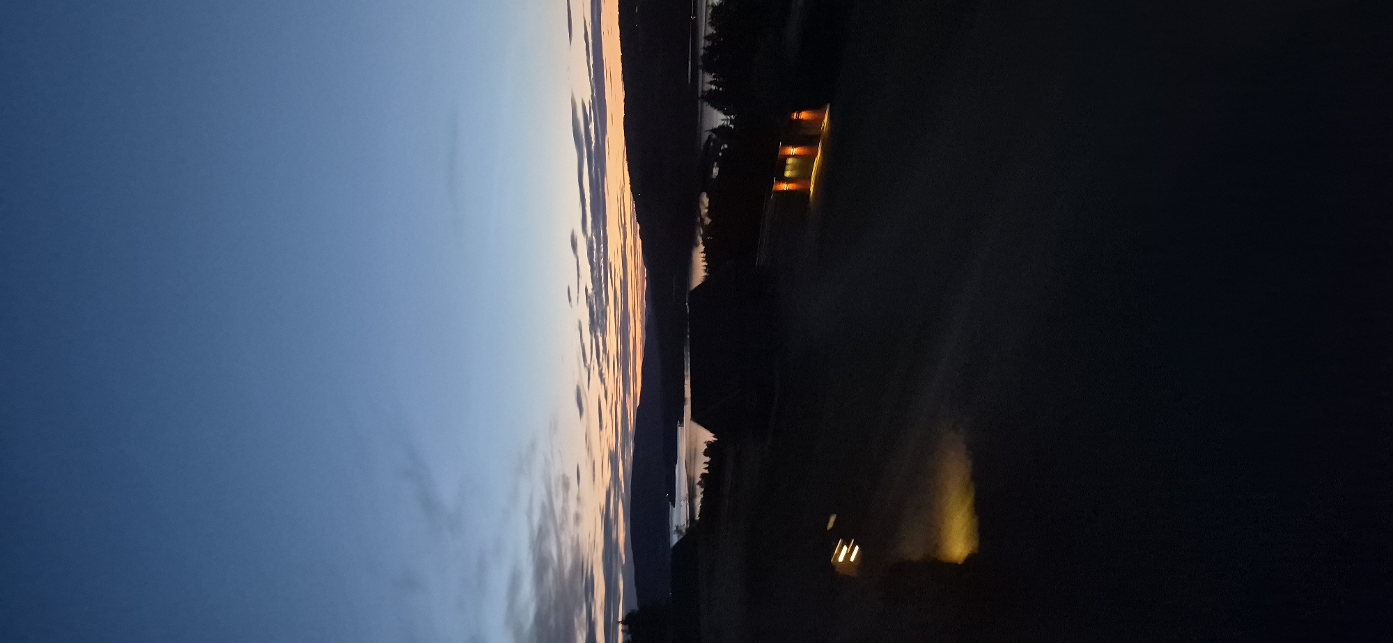The image depicts a twilight scene over a body of water, likely Lake Mjøsa, near Biristrand, Norway. The sky is a gradient of deep blue at the top, transitioning to lighter blues and then to hues of orange and pink near the horizon, indicating the setting or rising sun. Wispy clouds are scattered across the sky.
In the foreground, the land is dark and silhouetted. Two dark structures, appearing to be buildings, are visible. One, closer to the center and slightly to the right, has several illuminated windows casting a warm, orange glow onto the surrounding dark ground. The other building is partially visible on the left, with a bright, yellowish light emanating from it, possibly from headlights or a floodlight, casting an elongated reflection on the ground. The ground itself appears to be a field or open area, also mostly in shadow.
In the distance, rolling hills or mountains form a dark silhouette against the brightly colored sky. The body of water stretches between these hills, reflecting the sky's colors and appearing as a band of lighter tones. Trees are sparsely scattered along the shoreline. The overall impression is one of a peaceful, serene evening in a rural or semi-rural setting.