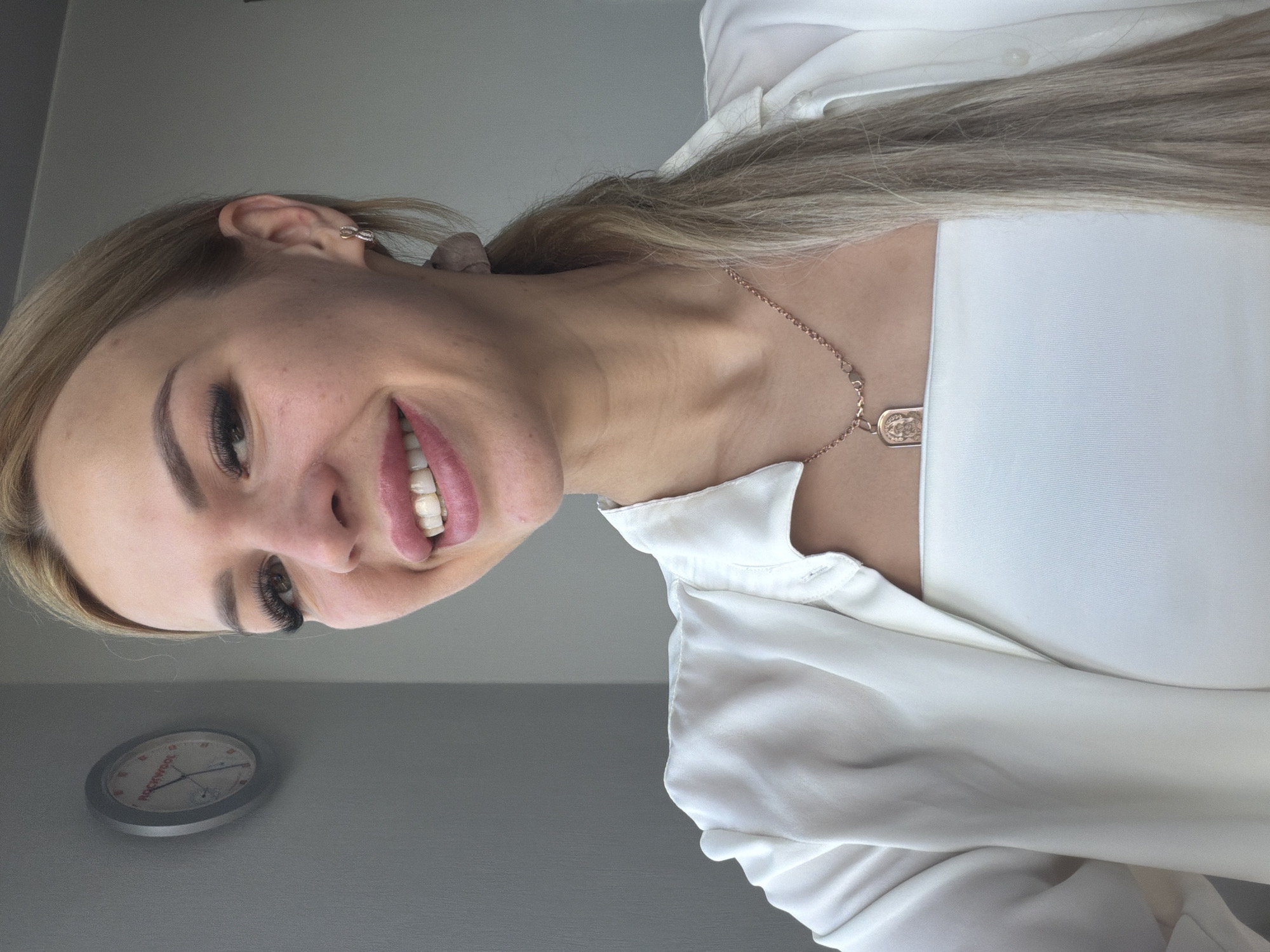 The image shows a close-up, waist-up shot of a young Caucasian woman with blonde hair tied in a ponytail, wearing a white sleeveless top and a white open shirt. She is also wearing a rose gold necklace with a pendant and small hoop earrings. She is smiling, and her makeup includes dramatic false eyelashes and pink lipstick. The background is a plain grey wall with a circular clock on it. The clock is silver with a white face and red markings. Based on the clock face, which appears to have the word "ROCKWOOL" on it, it is likely an office or commercial setting. The lighting suggests an indoor environment, possibly near a window. There are no overt signs of the city of Kyiv, Ukraine, within the frame of the image.