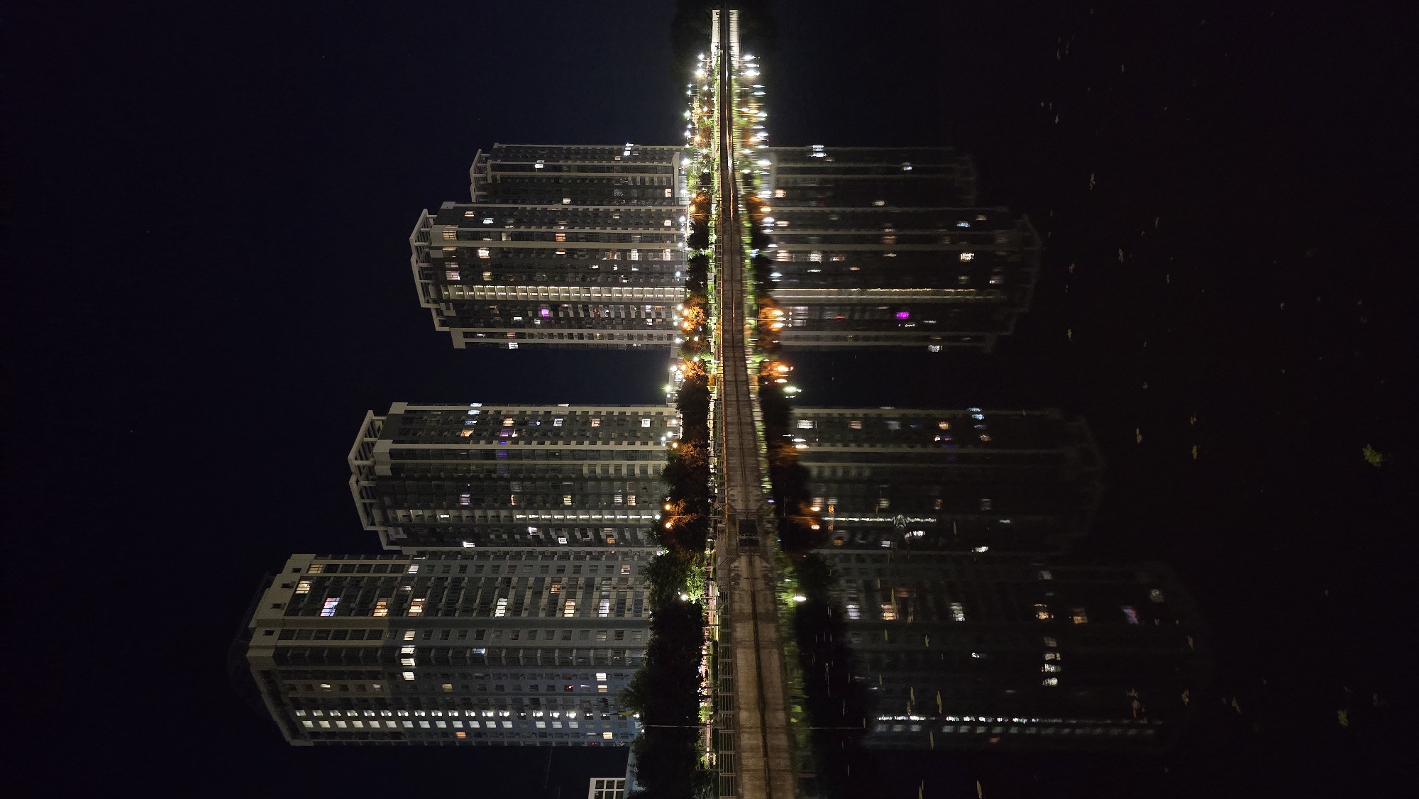 The image captures a nighttime view of several modern, multi-story apartment buildings on the banks of a body of water, likely a river or canal, in Kyiv, Ukraine. The buildings are illuminated, with lights visible in many of the windows, suggesting occupancy. They are tall and rectangular, with a consistent architectural style characterized by numerous windows and vertical architectural elements.
Along the waterfront, a paved promenade or walkway is visible, lined with trees and streetlights that cast a warm glow. The water in the foreground perfectly reflects the illuminated buildings and the lights from the promenade, creating a symmetrical and serene scene. The water's surface is dark, allowing the reflections to be sharp and distinct, though some scattered leaves or debris are visible on the surface. The sky is dark, indicating it is nighttime, with a few faint stars visible. The overall impression is one of urban development by the water, with a sense of calm and order.