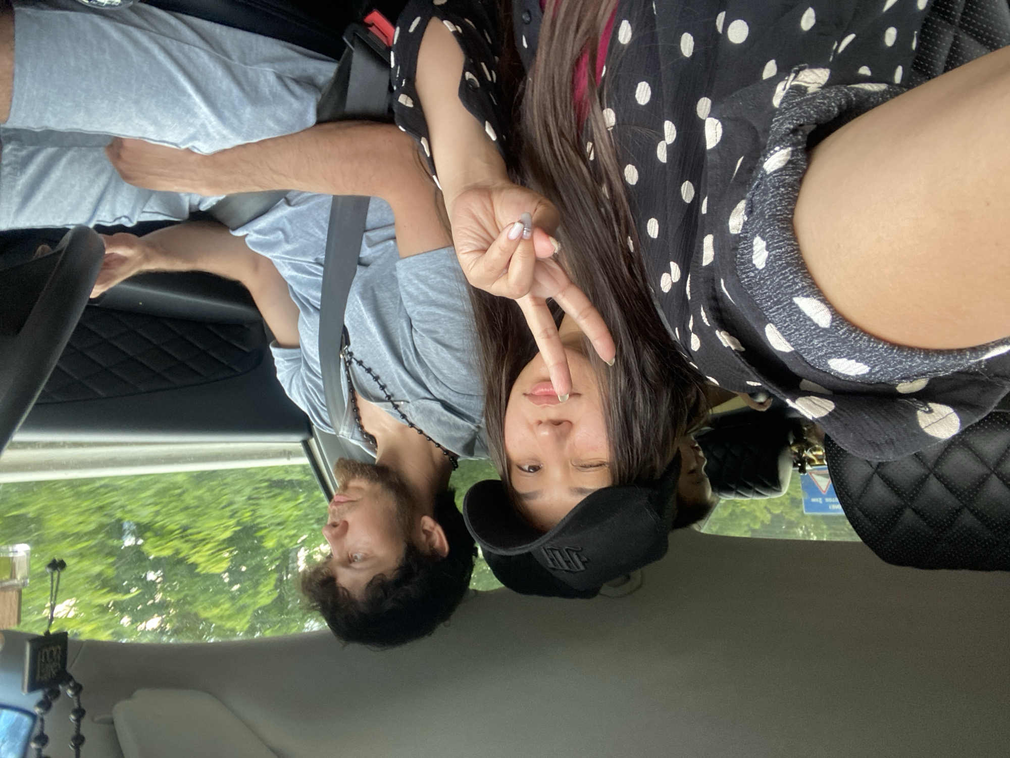 This is a point-of-view shot from inside a car, likely on a highway given the blurred trees and guardrail visible through the windows. In the foreground, a woman is leaning into the frame, making a peace sign with her right hand. She is wearing a black cap and a black and white polka dot shirt. To her right, a man is seated in the driver's seat, looking forward and gripping the steering wheel. He is wearing a light blue t-shirt and pants, with a beaded necklace. A child is partially visible in the background, between the two adults. The car's interior is visible, including a quilted seatback and a rearview mirror with a hanging ornament. The lighting suggests it is daytime. The image provides no explicit information placing it in Zilair, Russia.
