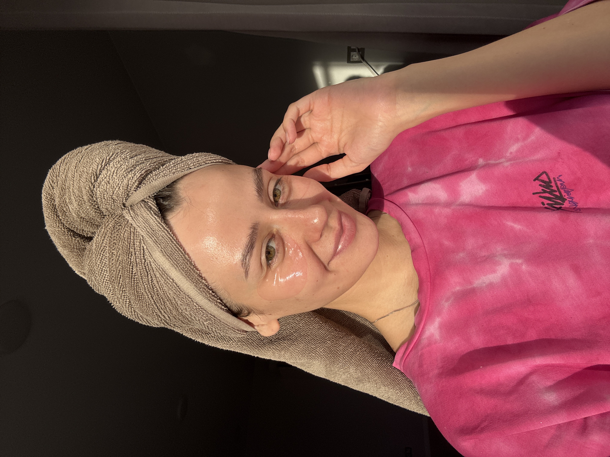 A person is photographed indoors with a beige towel wrapped around their head. Two clear gel patches are placed under their eyes. The person is wearing a bright pink, tie-dye shirt. The background is dark. No evidence suggests the location is Krasnoyarsk, Russia.