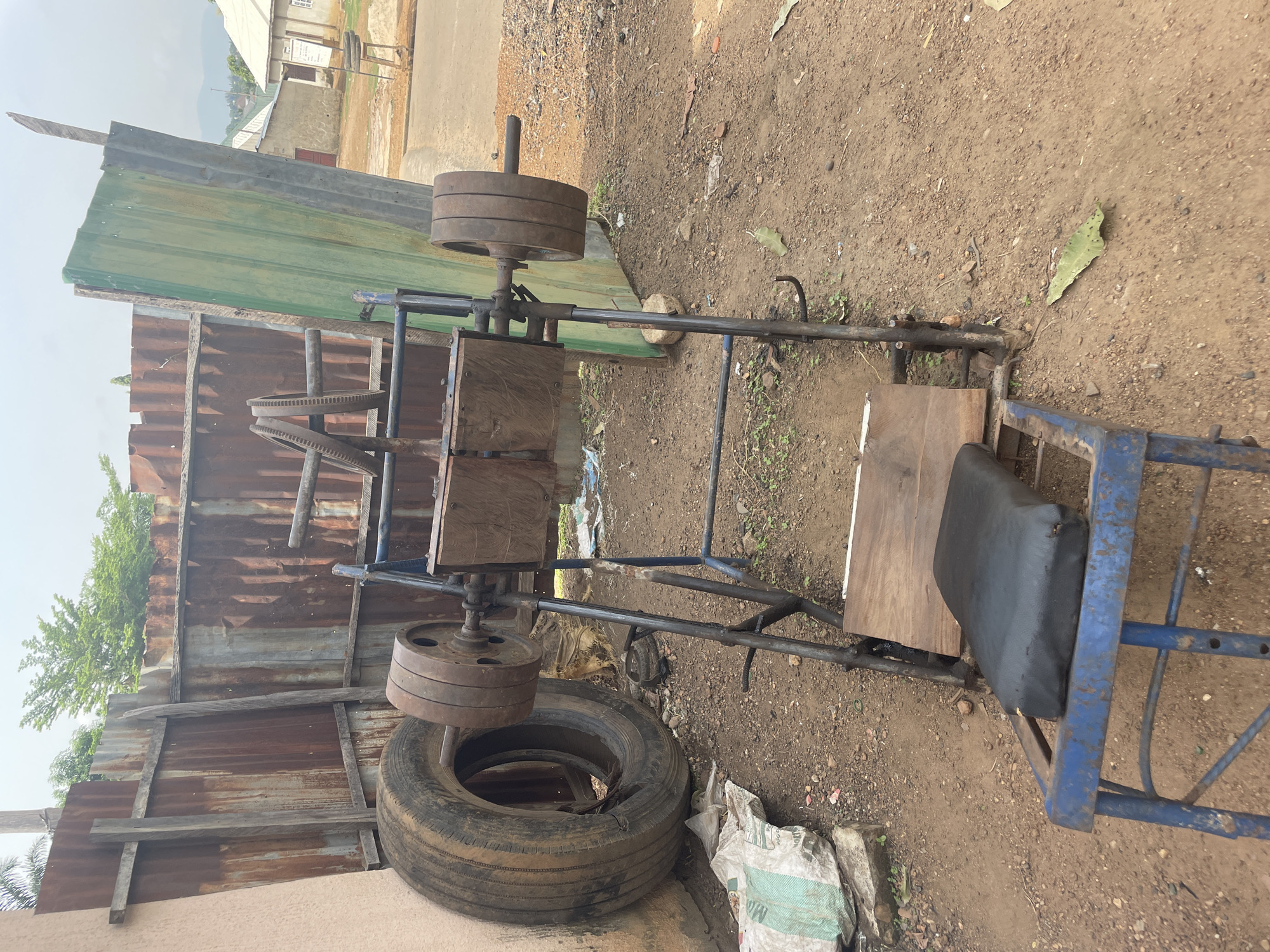 An outdoor scene in Akwanga, Nigeria, features a homemade weightlifting machine, likely a leg press, fabricated from salvaged materials. The machine is predominantly blue-painted metal tubing, forming a sturdy frame. A black cushioned seat, affixed to a wooden base, is part of the user's station. Wooden planks are also incorporated into the design, possibly as platforms or support structures for the movement mechanism.
The resistance is provided by a stack of circular, rusty metal weight plates, positioned on a vertical bar at one end of the machine. Another stack of similar weights is visible near the base, resting on what appears to be part of the machine's lever system. A large, worn-out tire lies on the dirt ground beneath one side of the machine, possibly used for stability or as a counterweight. Adjacent to the tire is a white sack with "M. A." and other illegible text visible.
The ground is unpaved, consisting of dry, light brown soil with scattered small rocks and sparse green vegetation. In the background, there are various corrugated metal fences – one section is green, and another is rusty brown – suggesting an informal boundary. Beyond these fences, simple single-story buildings with light-colored walls and dark roofs are visible under a clear, bright sky. The overall impression is of a sunny day. No people are present, and no specific activity is taking place, but the setup clearly indicates an area for physical exercise or a workshop where such equipment is made or used.