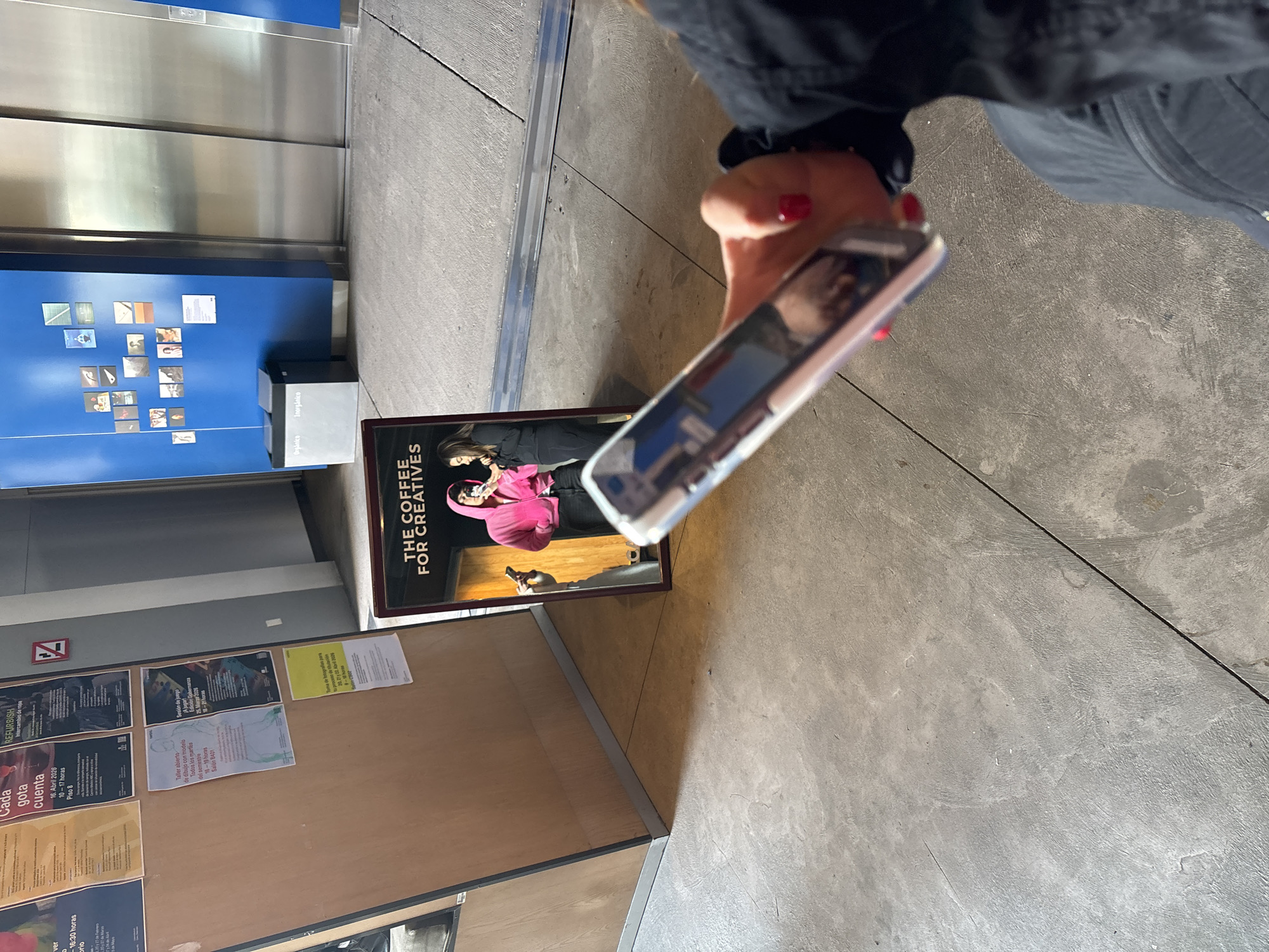 A person is holding a smartphone, appearing to capture a photo of a poster that reads "THE COFFEE FOR CREATIVES" and features a person in a pink outfit. The poster is positioned against a wall with other notices and flyers. Beside the poster is a metal elevator and a recycling station. The scene is in Mexico City, Mexico. The floor is grey concrete. The person holding the smartphone is wearing dark clothing.