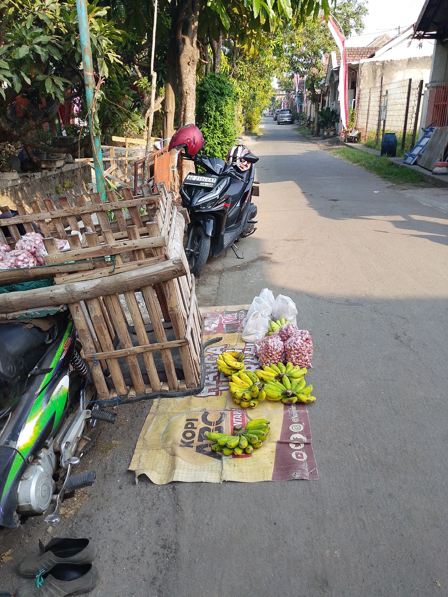 This is a daylight, eye-level, full-shot photograph of a street scene in Kediri City, Indonesia. In the foreground, a motorcycle is parked on the left side of the frame. Next to it, a wooden market stall displays produce, including bags of onions and bunches of bananas. A helmet rests on the motorcycle's handlebars. The street extends into the background, lined with houses and fencing. The weather appears clear and sunny, casting shadows on the asphalt road.
