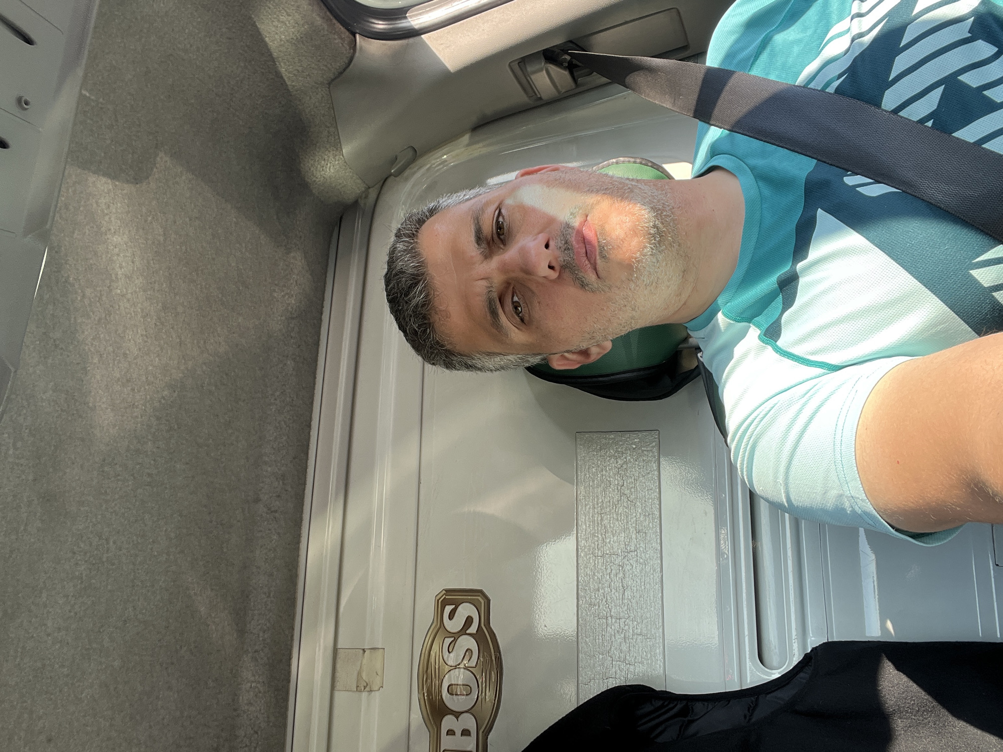 A man is taking a selfie inside a vehicle. He has short, graying hair and a goatee. He is wearing a teal and white t-shirt and a black seatbelt. The interior of the vehicle is visible behind him, including the ceiling and a portion of the driver's side door. There is a sticker on the wall of the vehicle that says "BOSS." The sun is shining through the window, casting shadows on the man's face and the interior of the vehicle. The image is taken from a slightly high angle, looking down at the man.
