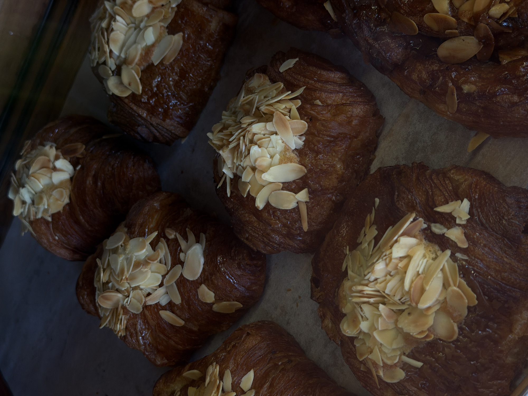 A close-up, overhead shot displays a collection of almond croissants. The croissants are golden brown and flaky, with a generous topping of thinly sliced almonds. Some of the croissants appear to be partially obscured by others. The lighting is dim, casting a warm glow on the pastries. The scene is likely set in a bakery or cafe on Koh Phangan, Thailand, given the city information.