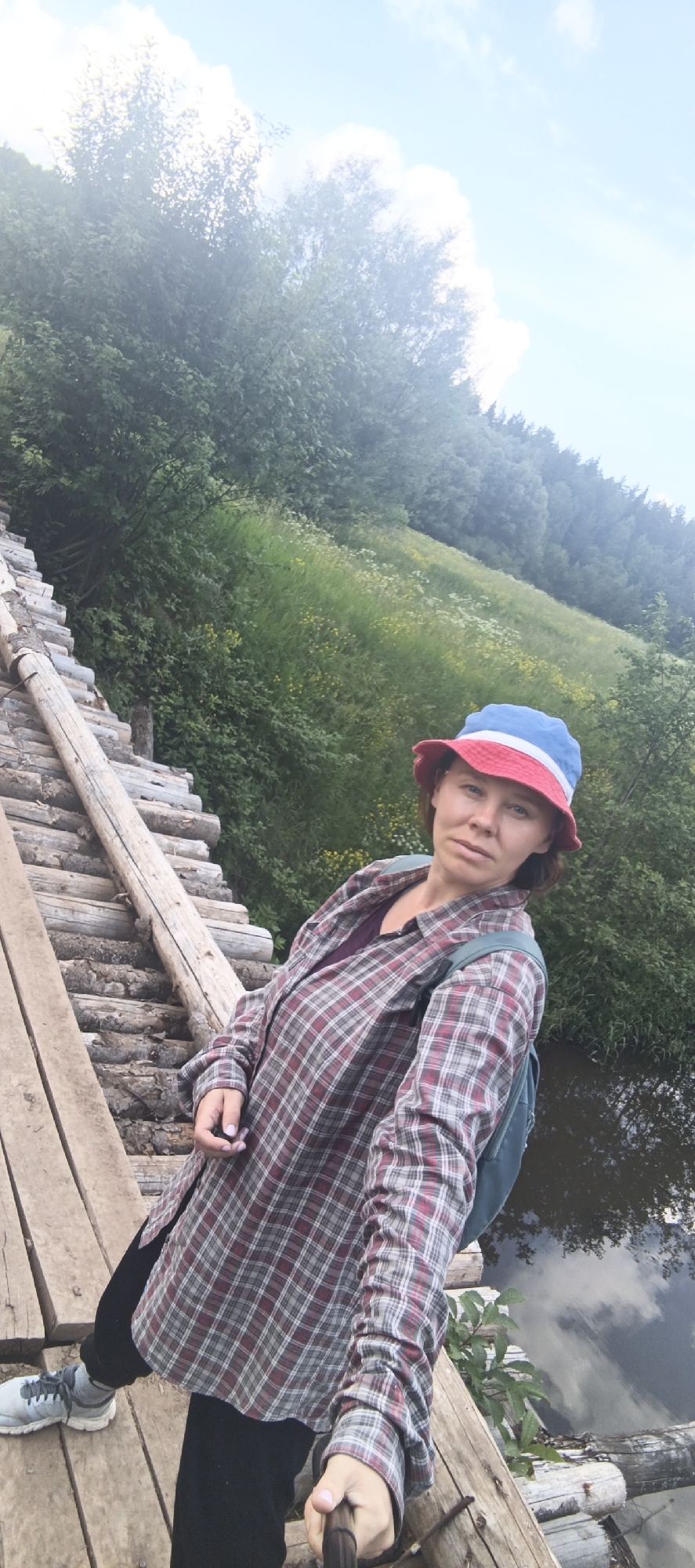 A woman is taking a selfie on a wooden bridge with a selfie stick. She is wearing a red, white, and blue bucket hat, a plaid shirt over a dark top, black pants, and grey sneakers. She has a backpack on. The bridge is made of logs and planks and crosses over a body of water with reflections of the sky. The background shows a grassy hillside with trees and some yellow wildflowers. The image appears to have been taken in Garevaya, Russia, based on the context provided.