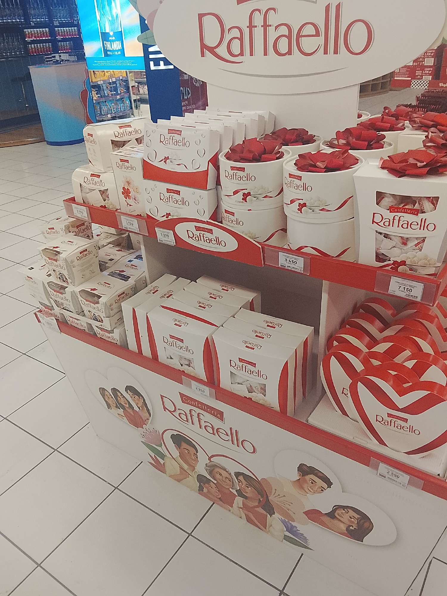 The image displays a confectionery product stand for "Raffaello" chocolates inside a retail store, located in an unknown city and country. The multi-tiered display unit is predominantly white with red accents, featuring the "Raffaello" logo prominently at the top. Numerous "Raffaello" products are arranged on the shelves, including white rectangular boxes, smaller square boxes, cylindrical containers topped with red crumpled decorative material resembling flowers or bows, and red and white striped heart-shaped gift boxes. Price tags are visible on the red shelf edges, indicating numerical values such as 3.170, 1.120, 1.370, 3.450, 7.150, and 2.590. The base of the display unit is adorned with graphic illustrations of smiling individuals of diverse ages and genders, suggesting themes of gifting and connection. In the background, to the left of the display, shelves stocked with bottles are visible, alongside a large digital screen advertising "FINLANDIA VODKA OF FINLAND" and a large blue and white striped cylindrical container. The floor is composed of white tiles with gray grout lines. No individuals are depicted interacting with the display in the immediate foreground.