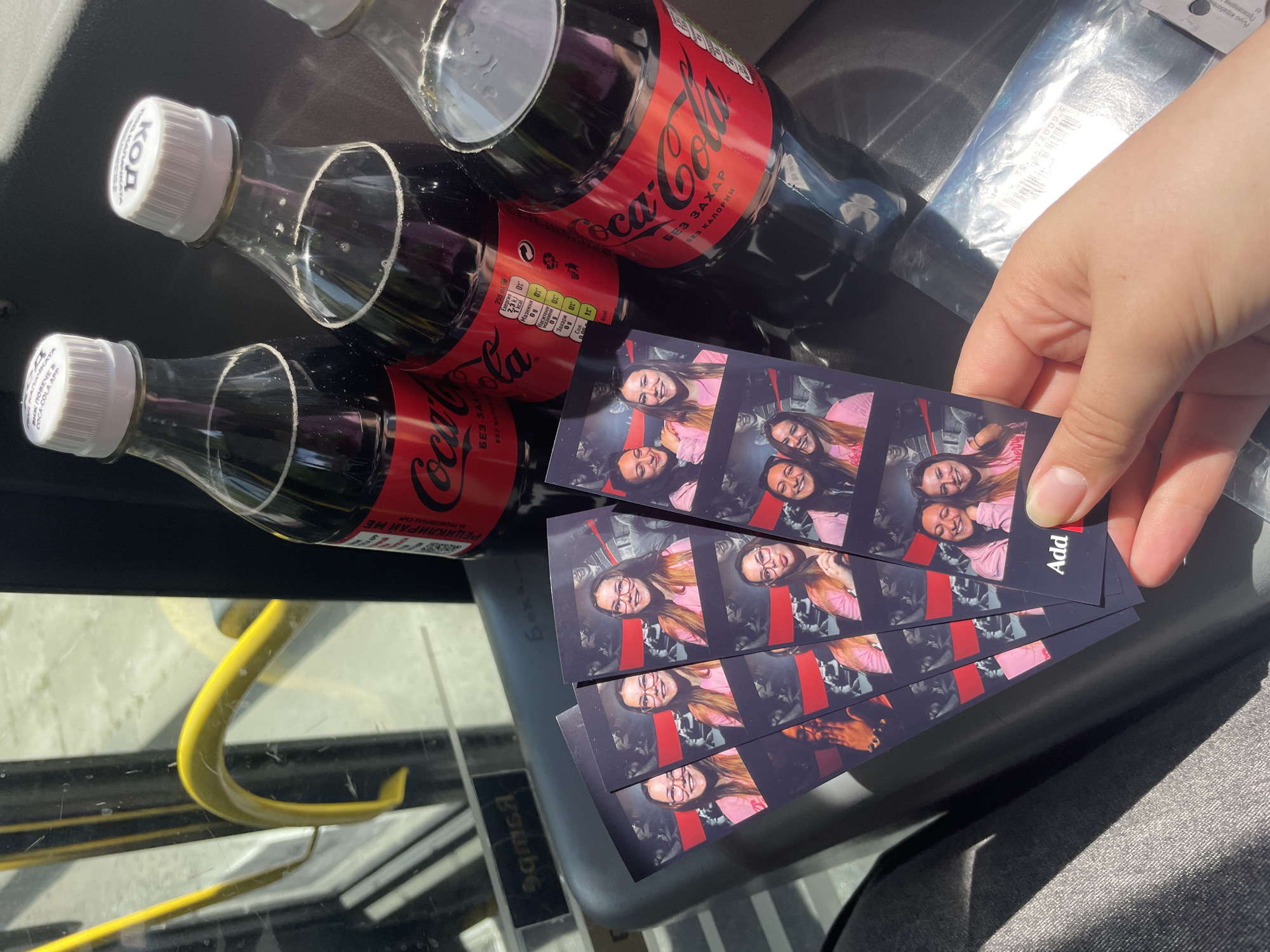 The image shows a close-up view of a hand holding several photo booth strips, next to three bottles of Coca-Cola, presumably in the city of Plovdiv, Bulgaria, based on the Cyrillic text on the Coca-Cola bottles and photo booth strips. The photo booth strips appear to feature two women in various poses. The Coca-Cola bottles are small, glass bottles with white caps. The background is somewhat indistinct, but it suggests an indoor setting, possibly a vehicle like a bus, given the presence of a yellow pole.