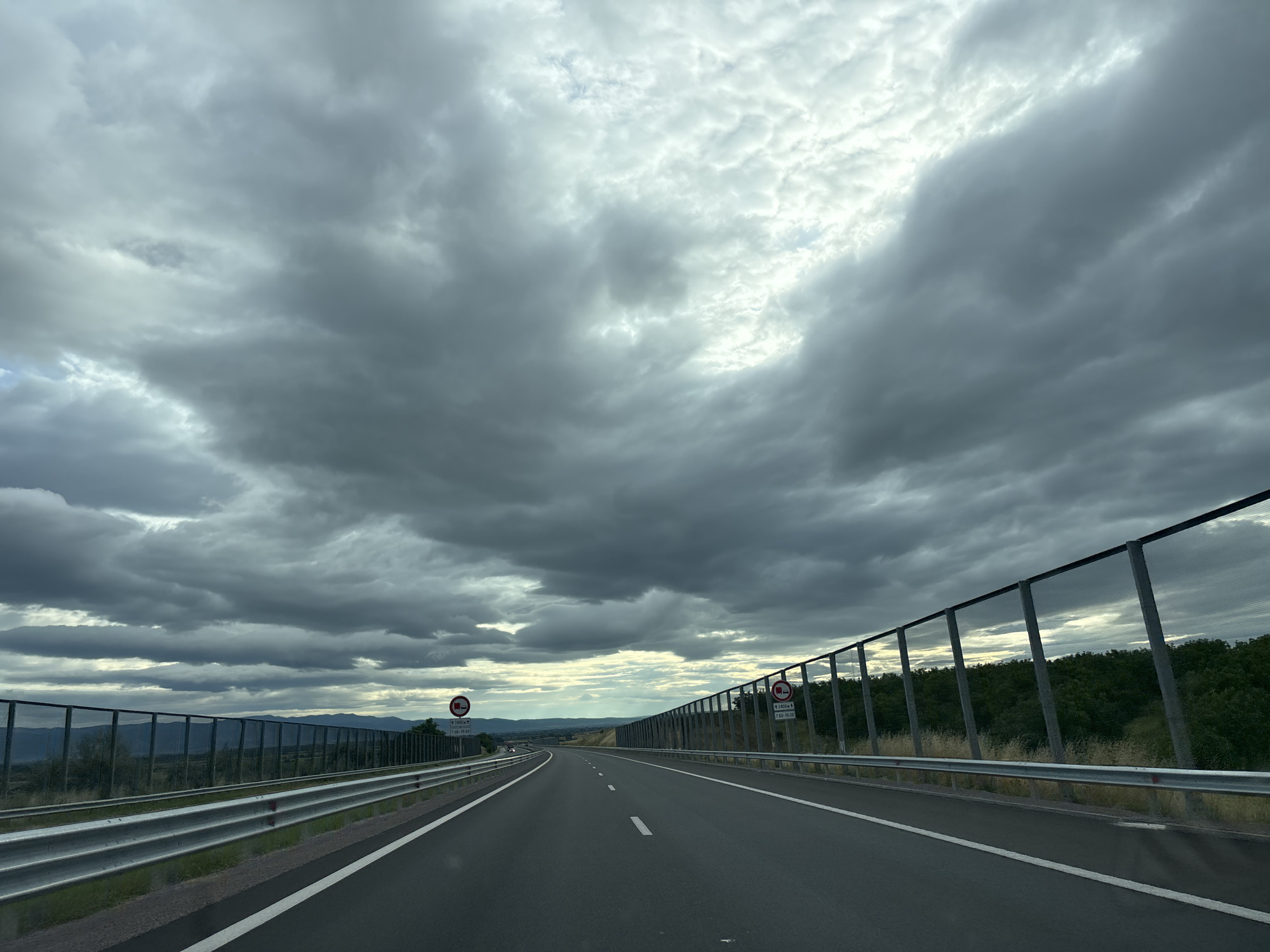 The image depicts a view from a car traveling on a highway. The road is asphalt with white dashed lines marking the lanes. On either side of the road, there are metal guardrails and fences. Beyond the fences, there are trees and hills. The sky is filled with dramatic, dark clouds, suggesting an impending storm or overcast weather. There are two traffic signs visible on the right side of the road, both displaying a red circle with a diagonal line, indicating a prohibition. One sign also has text below it. The location is identified as the city of Chokoba, Bulgaria. There are no people or animals visible in the image. The scene is captured from a moving vehicle, indicated by the slight blur of the surroundings.