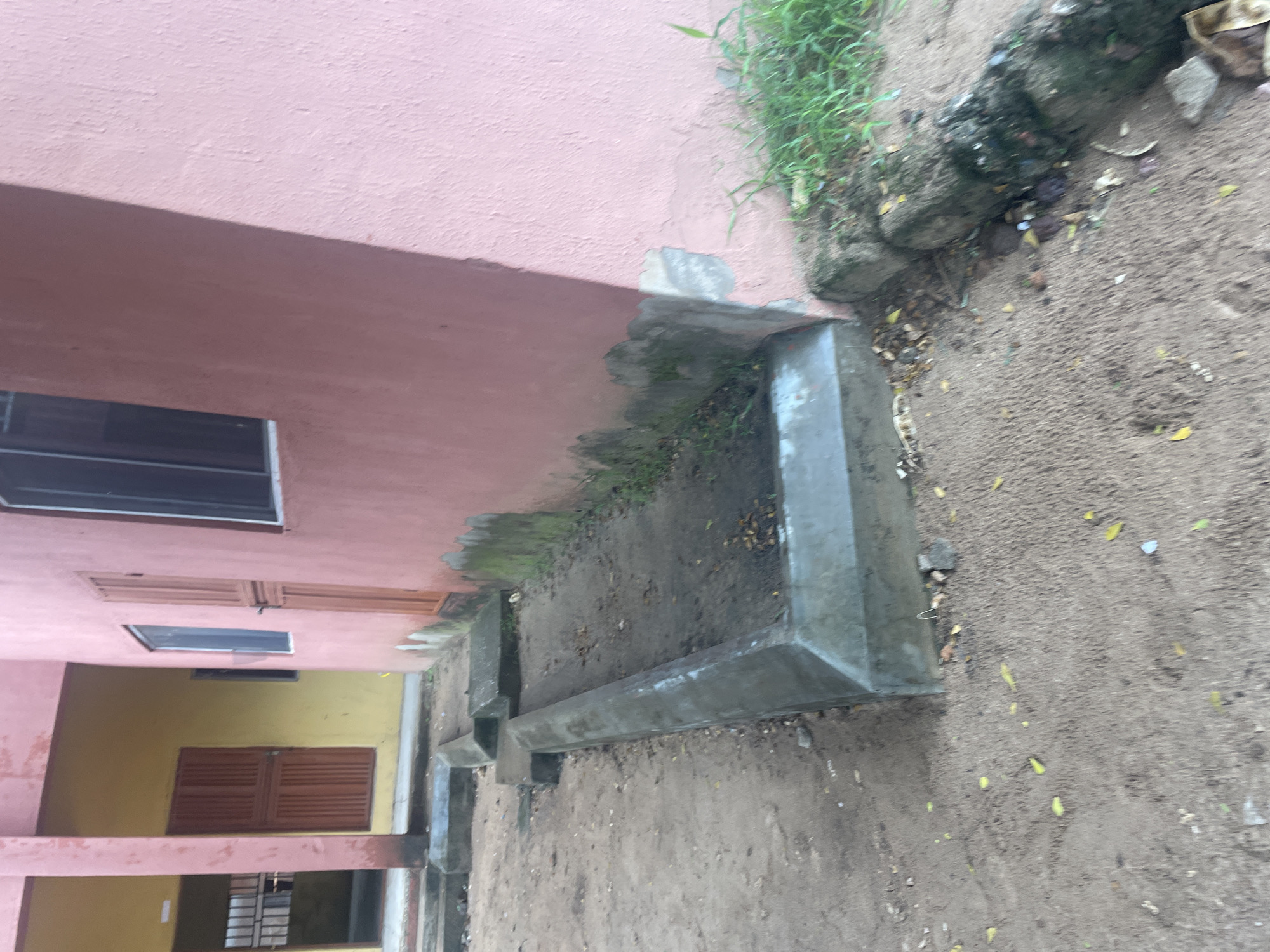 The image depicts an outdoor scene in Lokoja, Nigeria, featuring part of a building with pink and yellow walls. In the foreground, a long, concrete, V-shaped structure lies on the sandy ground. This structure appears to be some form of drainage or planter. The ground around it is covered in sand, with scattered leaves and debris. To the right, a section of the pink building shows signs of wear, with peeling paint and moss growth at the base. Some green grass is growing near the building. The building has at least one window visible and a brown door. The overall impression is of an outdoor area adjacent to an older building, possibly in a semi-urban or neglected setting.
