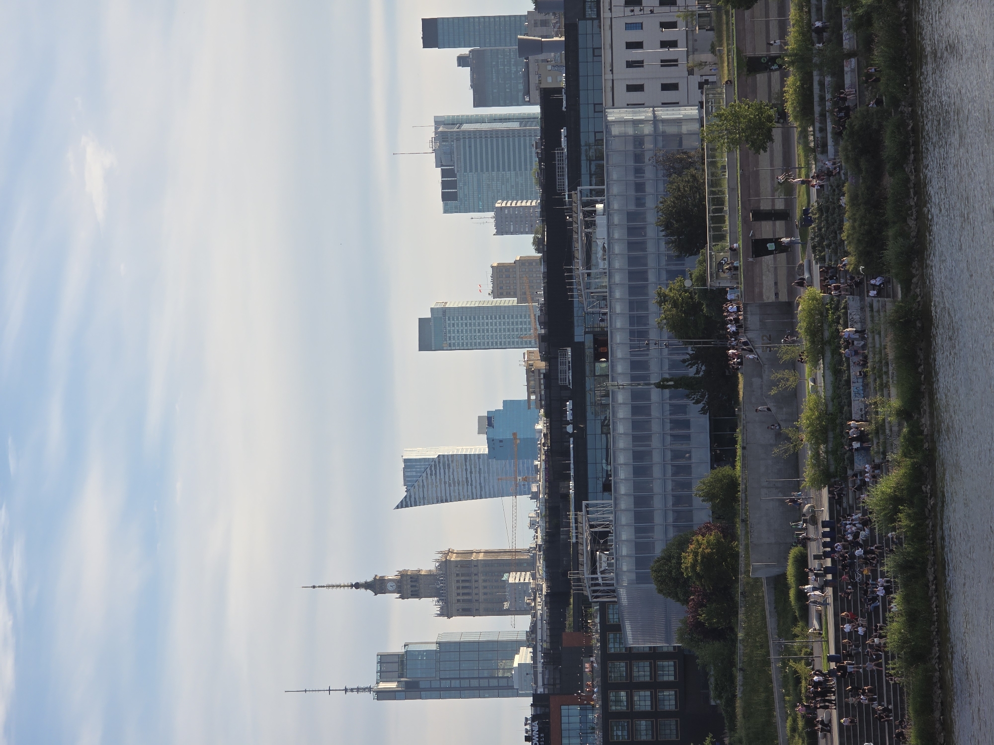 This is a wide shot of the Vistula River embankment in Warsaw, Poland, on a cloudy day. In the background, a modern cityscape of tall glass skyscrapers and older buildings, including the Palace of Culture and Science with its distinct clock tower, dominates the skyline. In the foreground, a tiered, amphitheater-like park area is filled with people of various ages relaxing, socializing, and enjoying the outdoors. Lush green vegetation and trees are interspersed throughout the park. The river in the very front is calm and reflects the sky and city.