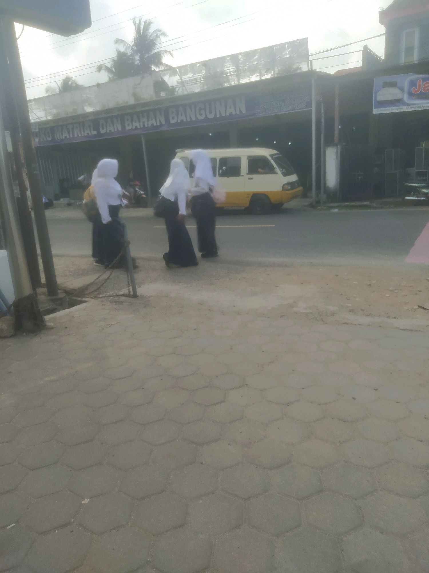 A street scene in Desa Karangbenda, Indonesia, features a yellow and white van parked on the right side of the road. Three young females in white hijabs and dark skirts stand on the sidewalk on the left side of the road. Behind them, a building with a sign that reads "TOKO MATRIAL DAN BAHAN BANGUNAN" (Hardware and Building Materials Store) is visible. The sidewalk is paved with hexagonal concrete blocks. Palm trees are visible in the background, suggesting a tropical climate. The lighting indicates it is daytime, possibly overcast.
