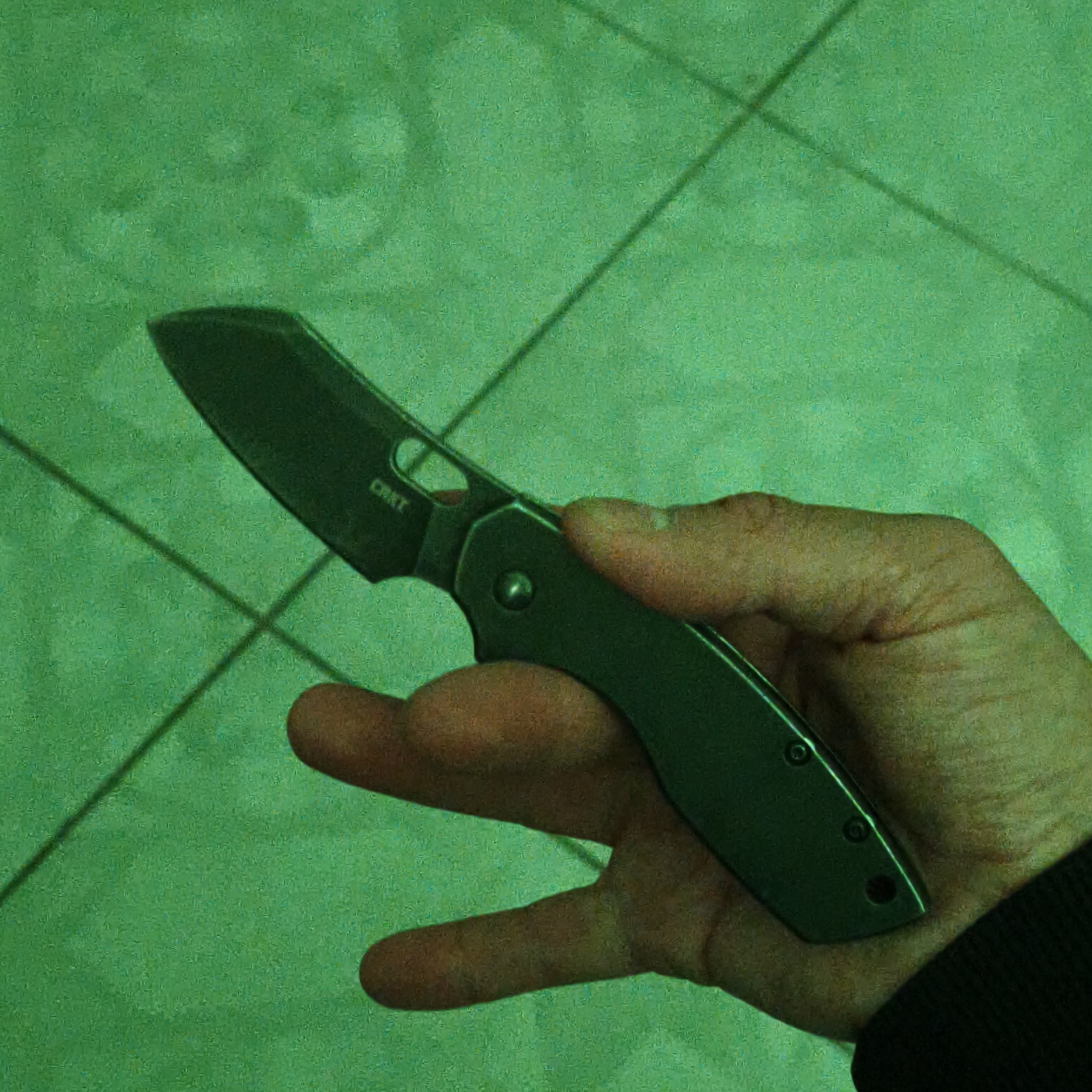 A person is holding a folding knife in their right hand. The knife appears to be green and has the brand "CRKT" visible on the blade. The background features a green tiled floor with lines suggesting a pattern. The green cast suggests the image was taken under non-natural lighting conditions. The setting does not provide enough information to accurately pinpoint a location in Hrodna, Belarus.