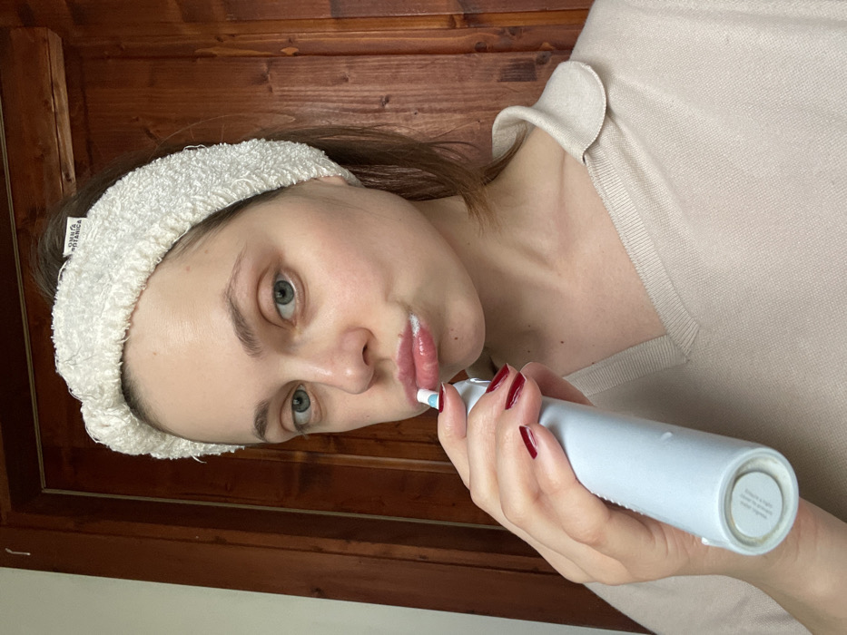 A forensic examination of the image reveals a close-up, chest-up portrait of a Caucasian female, appearing to be in her 20s or 30s, engaged in a personal hygiene routine within an indoor setting in Monsummano Terme, Italy. The subject has fair skin, light-colored eyes, and light brown hair pulled back, secured by a white, textured terry-cloth headband displaying a small black tag with "OMNIA BOTANICA" visible on the right side of her head. She is wearing a light beige or cream V-neck garment with a subtle collar. Her right hand, adorned with dark red nail polish, holds a light blue electric toothbrush with a white and blue brush head to her partially open mouth. White toothpaste foam is visible around her lips. The subject is looking directly into the camera. The background consists of a dark brown wooden surface, likely a door or wall paneling, with visible wood grain.