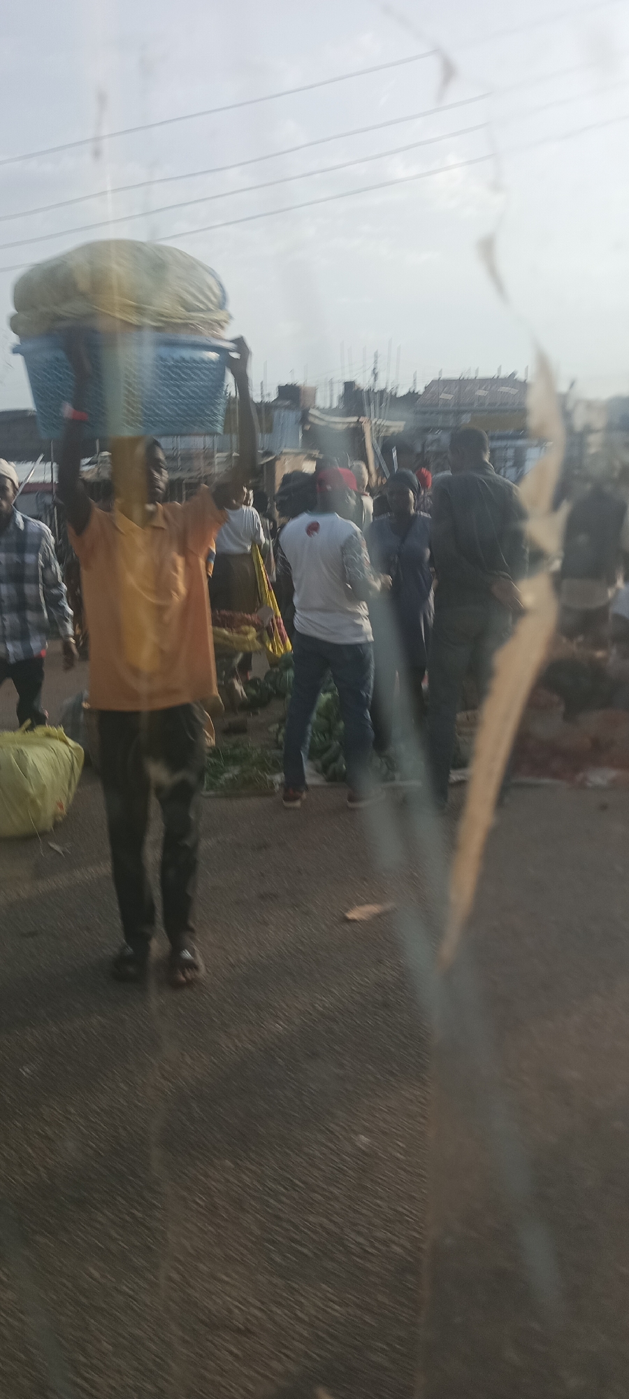 The image depicts a bustling outdoor market or street scene in Gura Top, Nigeria. Numerous people are visible, engaged in various activities, suggesting daily commerce and movement.
In the foreground, a man wearing an orange t-shirt and dark pants stands, expertly balancing a large, light blue woven basket filled with light-colored contents (possibly produce or textiles) on his head. He wears flip-flops. To his left, a yellow sack rests on the ground. Other individuals are scattered throughout the mid-ground and background. One man wears a white t-shirt with patterned sleeves and blue jeans, while another is seen from behind in dark attire. Piles of green produce, possibly watermelons or cabbages, are arranged on the ground in the mid-right.
The scene is set on what appears to be an asphalted or dirt road. In the background, numerous informal structures and buildings are visible, indicative of an urban or semi-urban environment. Overhead power lines stretch across the sky, which is hazy or overcast, suggesting a daytime shot with diffused light, possibly early morning or late afternoon. The overall mood is functional and busy. No legible text is visible in the image. The photograph appears to have been taken through a window, causing a blurred streak on the right side.