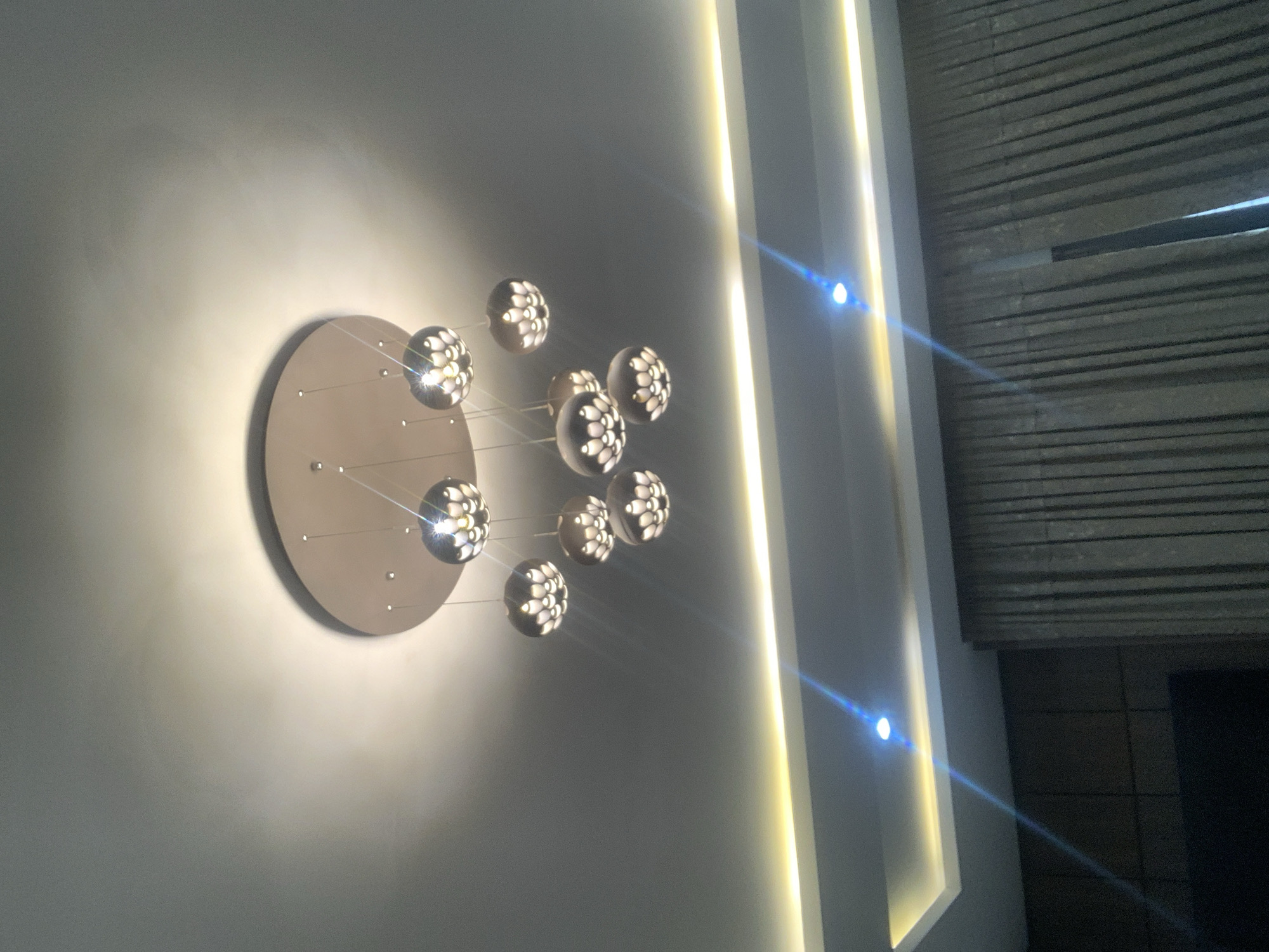 The image shows a modern interior with a chandelier and recessed lighting. The chandelier features a circular ceiling plate from which several spherical lamps hang at varying lengths. The lamps have an intricate design with multiple openings that emit a soft glow, resembling a floral pattern. Below the chandelier, recessed lighting fixtures are visible along a dropped ceiling, with one emitting a blueish light streak due to lens flare. Dark, textured curtains hang on the right side of the image, and a dark paneling is visible on the left. The overall impression is one of contemporary, elegant home decor. The scene is consistent with an indoor setting in a residential or commercial property, and such modern lighting designs are common in upscale properties in Abuja, Nigeria.