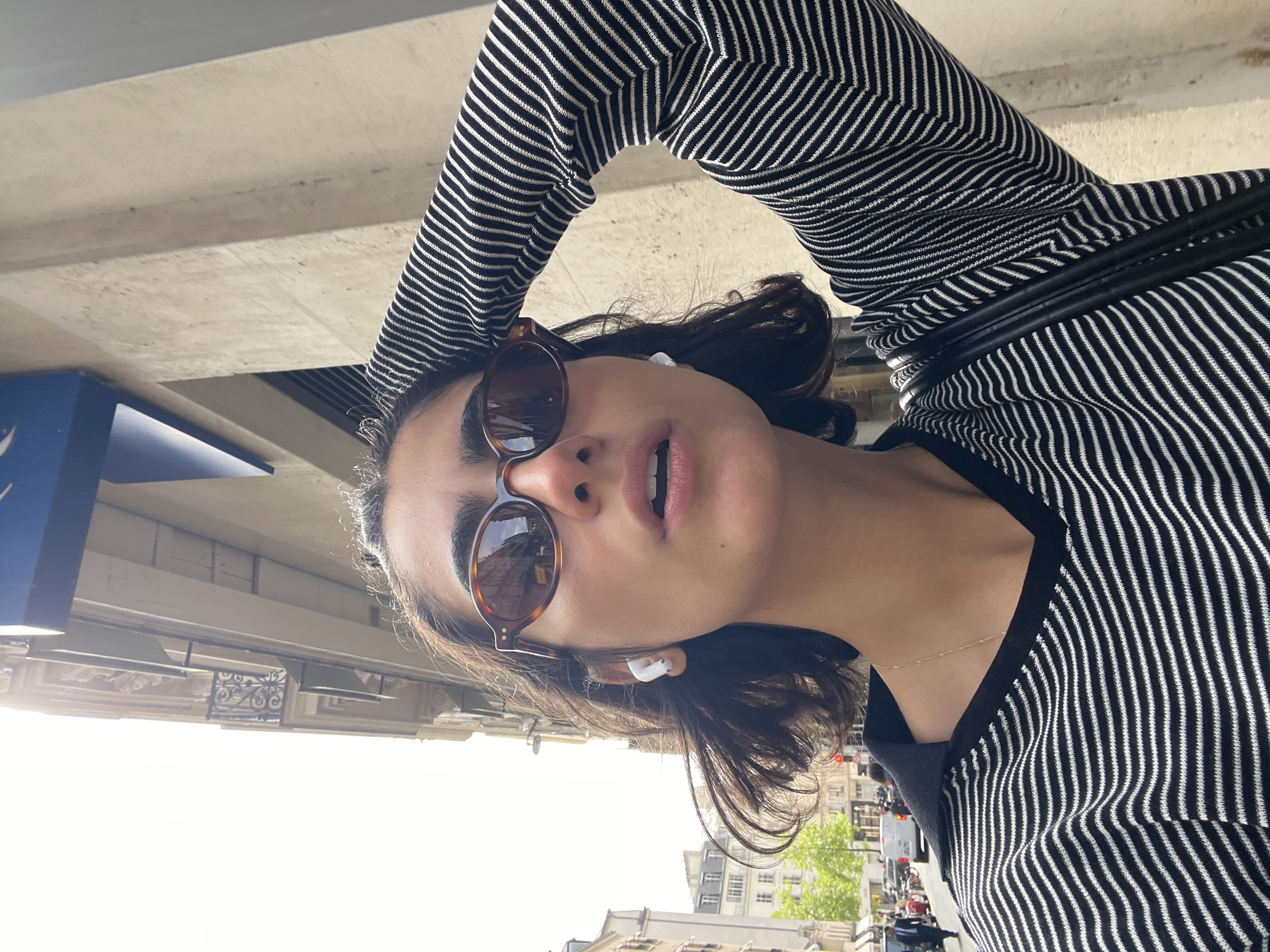 This image is a selfie taken from a low angle, rotated 90 degrees clockwise, depicting a person outdoors in Paris, France. The subject, likely female, has dark hair and is wearing a black and white striped long-sleeved top. She is wearing round, tortoise-shell sunglasses and white wireless earbuds in both ears. A thin gold chain necklace is visible around her neck. Her mouth is slightly open, and she is looking directly towards the camera. A black strap, possibly from a bag, is seen draped over her shoulder.
The setting is a sunny urban streetscape. The background features light-colored, multi-story buildings typical of Parisian architecture, some with decorative elements and a wrought-iron balcony. Green foliage from trees lines the street, and distant glimpses of vehicles and other pedestrians suggest a lively city environment. The bright, clear sky indicates strong daylight, likely mid-day. A large concrete structure is visible overhead in the top left, along with a portion of a blue sign.
The activity captured is a casual moment, possibly a self-portrait taken while walking or pausing on the street. No discernible text is visible within the frame. The overall atmosphere is bright and energetic, reflecting a moment in a vibrant European city.