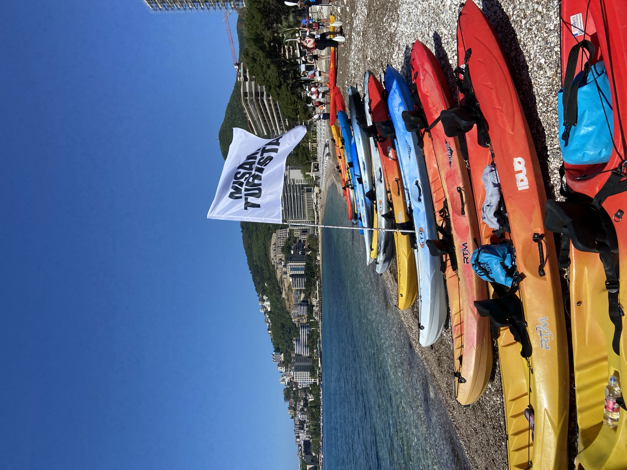 A sideways outdoor shot shows a row of kayaks lined up on a pebble beach in Budva, Montenegro. The kayaks are various colors including red, orange, blue, yellow, and white. Several are labeled with "RTM". A white flag with black writing that says "MISAD TURISTA" is planted in the beach. In the background is the city of Budva, with mountains rising behind the city. The sky is clear blue. Some people are standing at the top of the image. The Adriatic Sea is visible in the middle ground. A building under construction with a crane is visible in the upper-left.