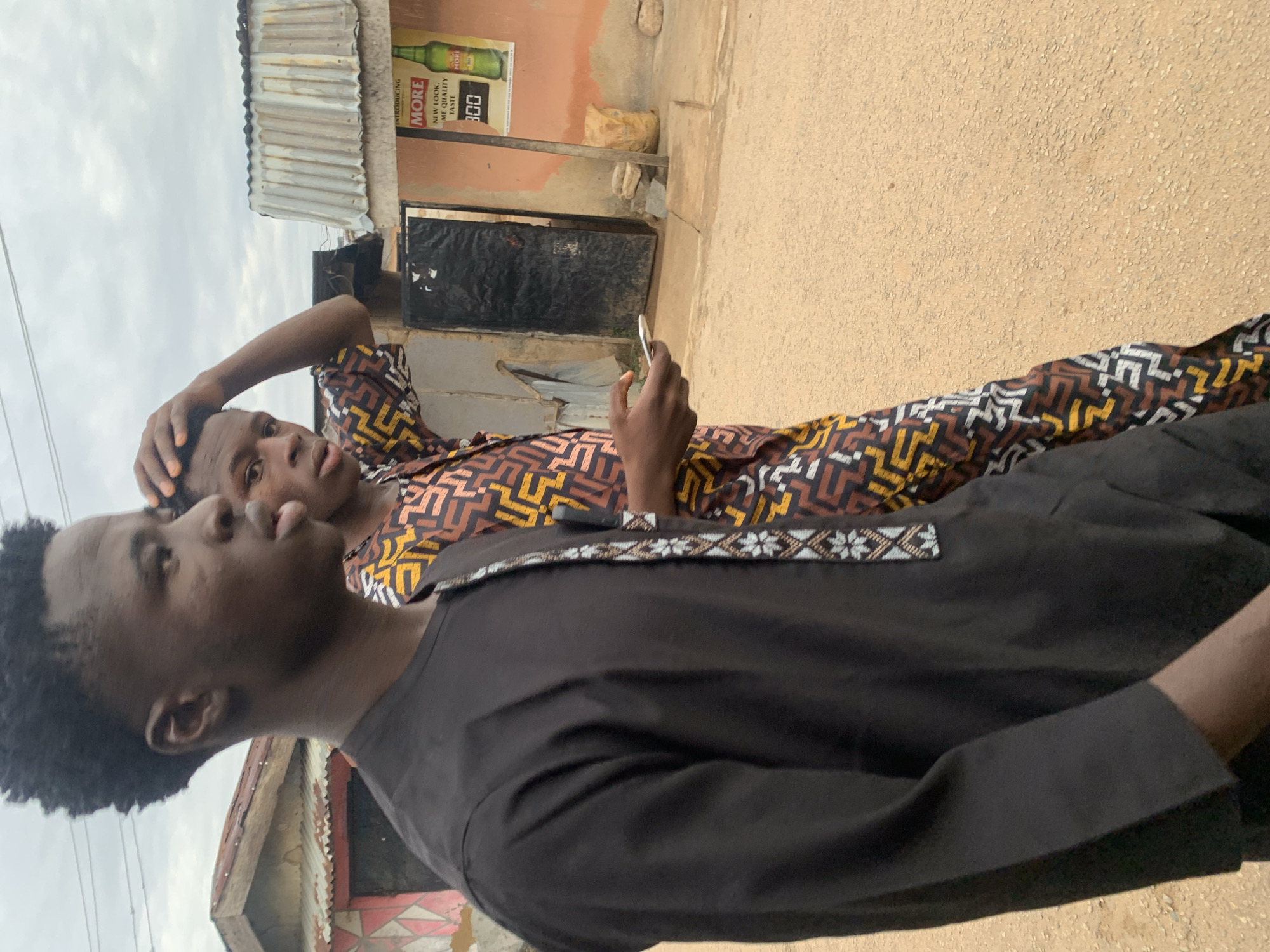 The image depicts two young Black males walking on an unpaved, light brown road under an overcast sky in what appears to be a residential or commercial area in Jos, Nigeria.
The male in the foreground, positioned in profile to the left, has short, dark, curly hair and a distinct hairline. He wears a long-sleeved black tunic featuring a vertical strip of light blue, white, and brown geometric patterns along the right side of the chest. His mouth is slightly open, and his right arm is bent with his hand visible.
The male behind him, to the right, has similar short, dark, curly hair and is looking towards the viewer with his mouth slightly open. He is wearing a long-sleeved shirt patterned with intricate geometric shapes in shades of brown, yellow, black, and white. His right hand is placed on his head, while his left hand holds a white smartphone. He wears dark footwear on his visible left foot.
In the background, to the left, a single-story building with a reddish-brown corrugated metal roof and a light-colored wall, featuring faint geometric patterns, is visible. Overhead wires are present against the cloudy sky. To the right, a light brown-painted wall displays a poster advertising "MORE" brand, which includes text such as "INTRODUCING MORE," "NEW LOOK, ME QUALITY TASTE," and "300," alongside an image of a green bottle. A wooden pole stands near the poster, and a light-colored sack with several rocks rests on the ground. Further back, a dark, open doorway is visible on a building.