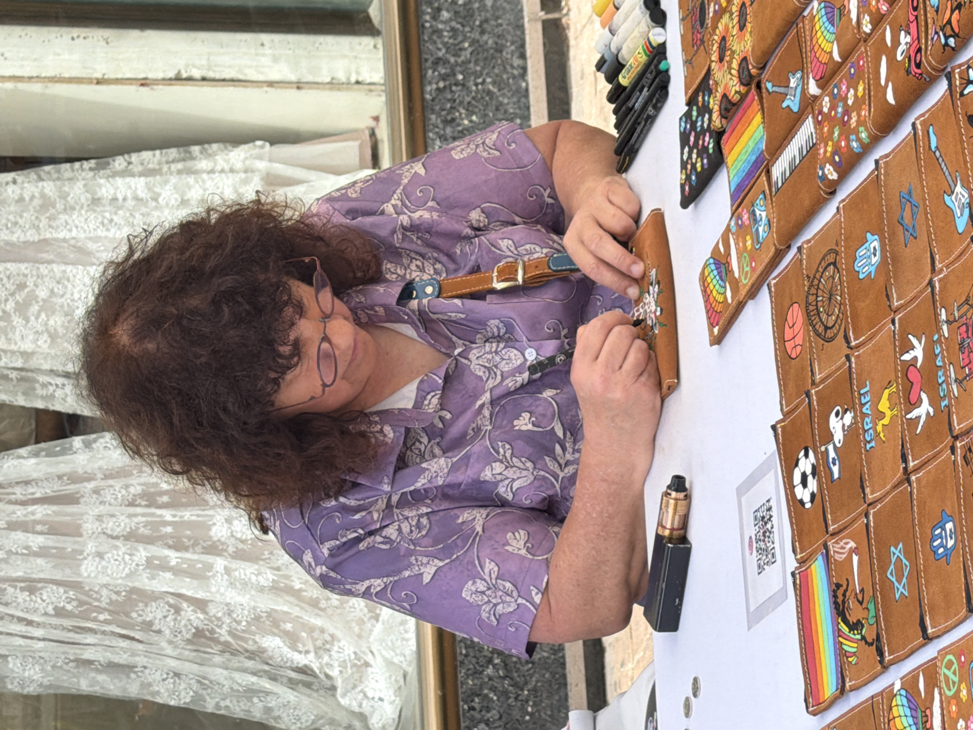 A woman with curly brown hair and glasses, wearing a purple floral shirt, is working at a table displaying brown leather wallets and purses with various colorful designs including sunflowers, rainbows, and symbols such as the Star of David and the Hamsa. She is adding detail to one of the wallets. Several markers and a vape pen are also on the table. A QR code in a white frame is visible on the table. The background suggests the edge of a building and a window with lace curtains. The image was most likely taken in the city of Tel Aviv, Israel, given the presence of wallets with "Israel" written on them.