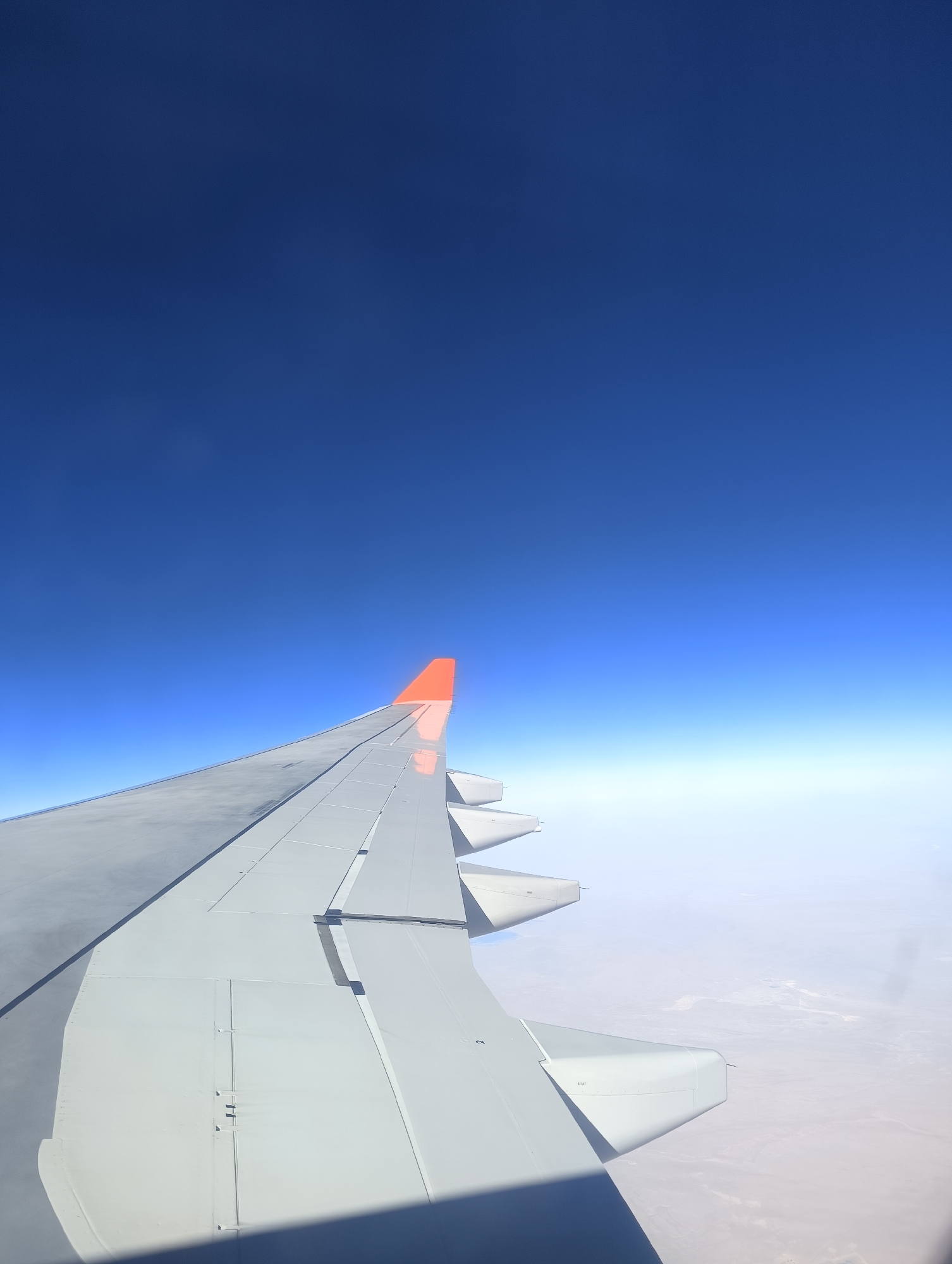 An aerial photograph captured from an airplane window shows a vast expanse of clear, deep blue sky above a hazy, light-colored horizon. The foreground is dominated by the wing of the aircraft, which is predominantly white or light gray. The wing has a sleek, aerodynamic design and features several panel lines and fasteners. The very tip of the wing is painted a bright orange, contrasting sharply with the rest of the wing and the sky. Below the hazy horizon, the terrain is visible as a pale, arid landscape, possibly desert or a dry plain, with faint hints of geological features. The overall impression is one of flight at high altitude over a remote and largely uninhabited area. The photograph does not depict the city of Nakhabino, Russia, as it shows a view from an aircraft in flight, not a ground-level perspective of a specific urban location.