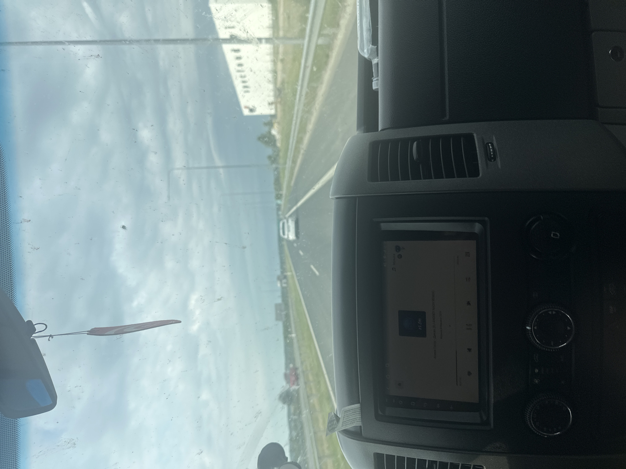 The image shows the interior of a vehicle driving on a highway. The dashboard is visible, with a car stereo displaying music information. A pink air freshener hangs from the rearview mirror. The windshield is dirty, with streaks and smudges. Outside the vehicle, the highway stretches ahead, with other cars visible in the distance. There are streetlights and buildings lining the road. The sky is cloudy and overcast. The location is stated to be Stawiski, Poland, though this cannot be visually confirmed from the image alone.